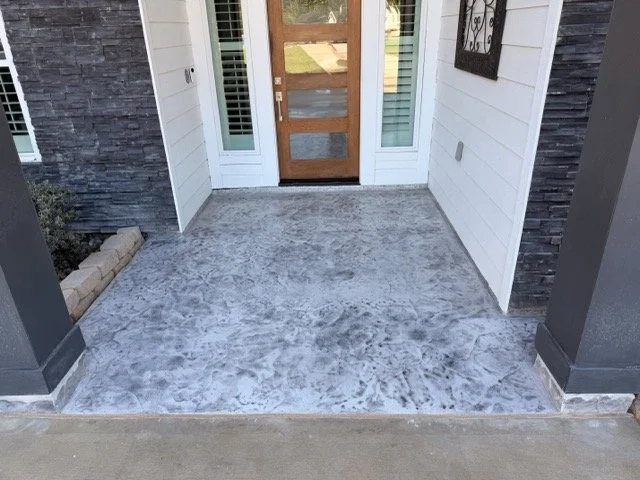 Front porch with textured concrete floor, wooden door with glass panels, white siding, and decorative wall hanging