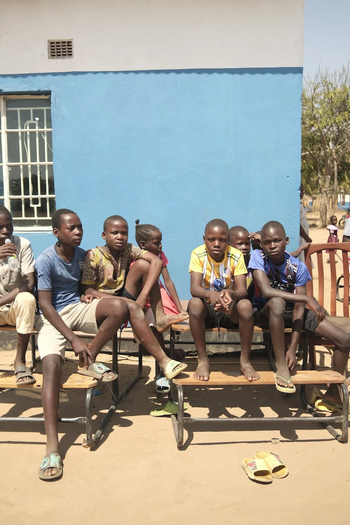 A group of young people sit on a bench wearing flip flops, t-shirts and shorts. The weather seems warm. There is sand underfoot, and they are sitting in front of a building with a blue wall.