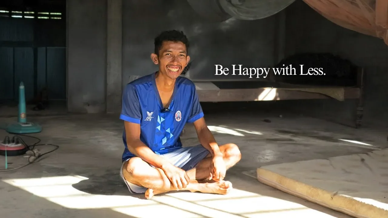 A man wearing a blue shirt sits on a concrete floor, with a large smile. The room around him seems empty, with just a bed in the background and a charging plug socket. Text overlaid on the image reads, 'Be Happy with Less'.