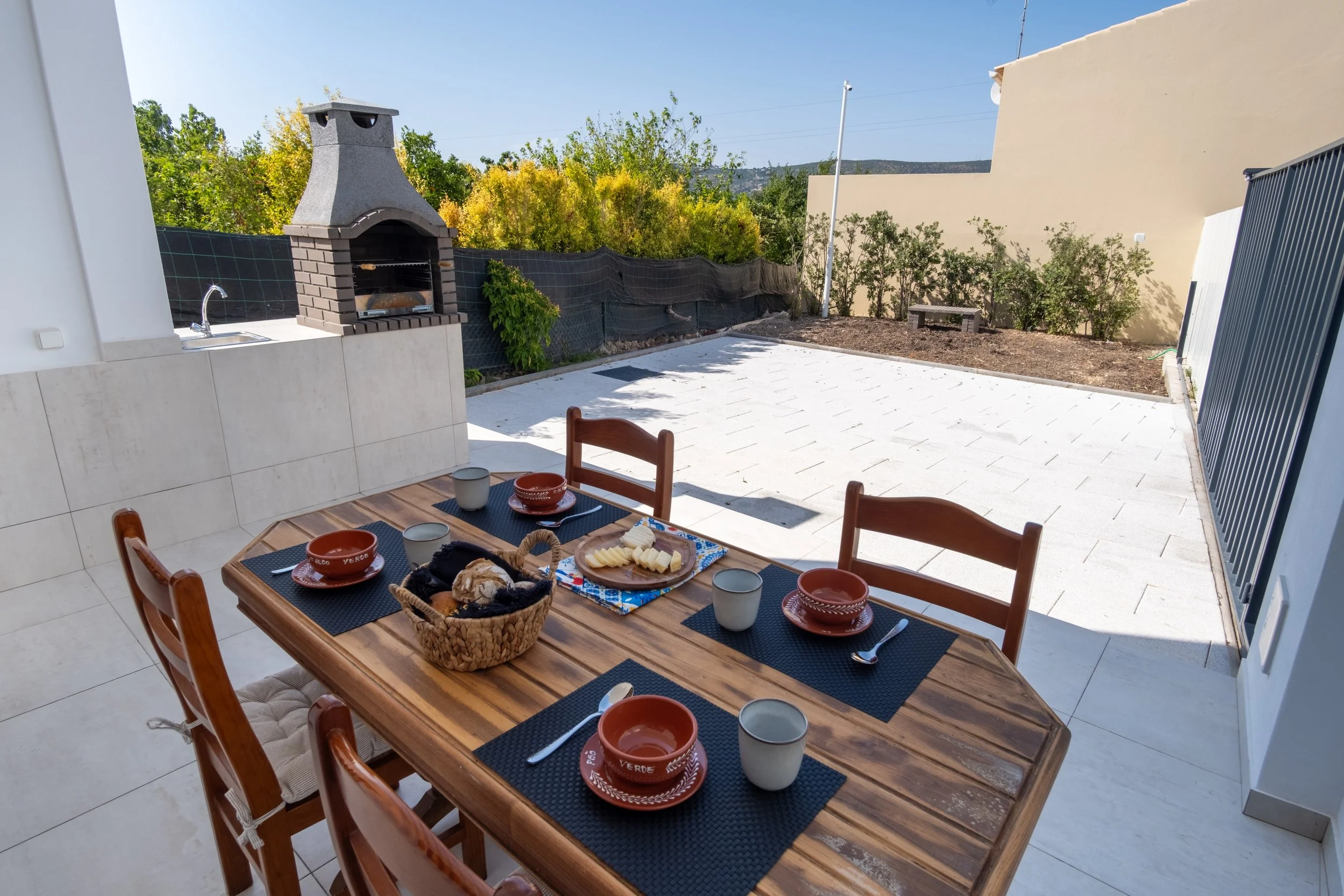 Mesa de jantar ao ar livre com pratos e utensílios, churrasqueira ao fundo em um espaço externo com piso de cerâmica e jardim com arbustos e árvores.