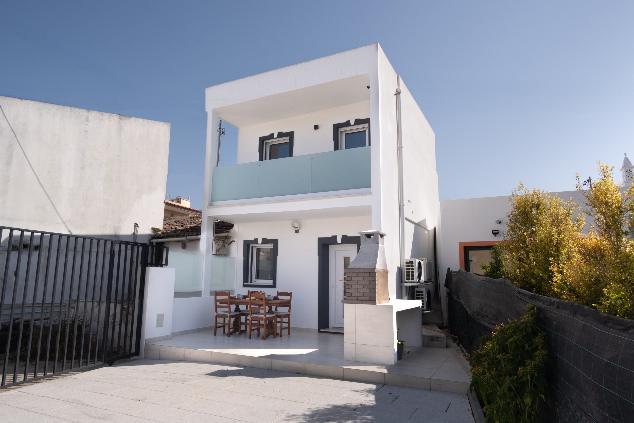 Casa moderna com varanda de vidro na parte superior, área externa com mesa de jantar e churrasqueira, cercada por muro e vegetação, sob céu claro.
