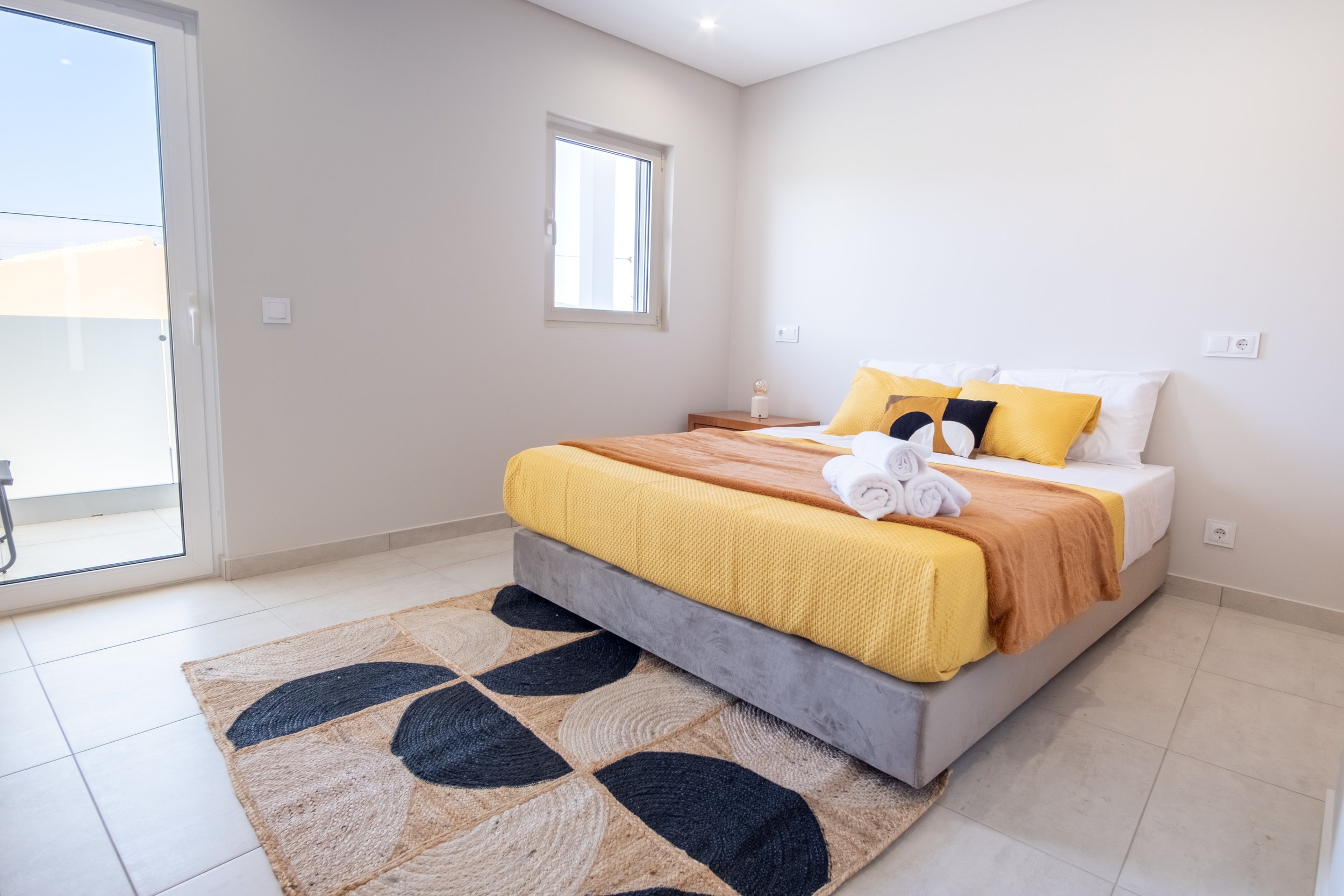 Quarto com cama de casal, roupa de cama amarela, almofadas amarelas e preta, toalhas brancas dobradas, janela, porta de vidro para varanda, tapete com padrão preto, bege e branco, parede branca