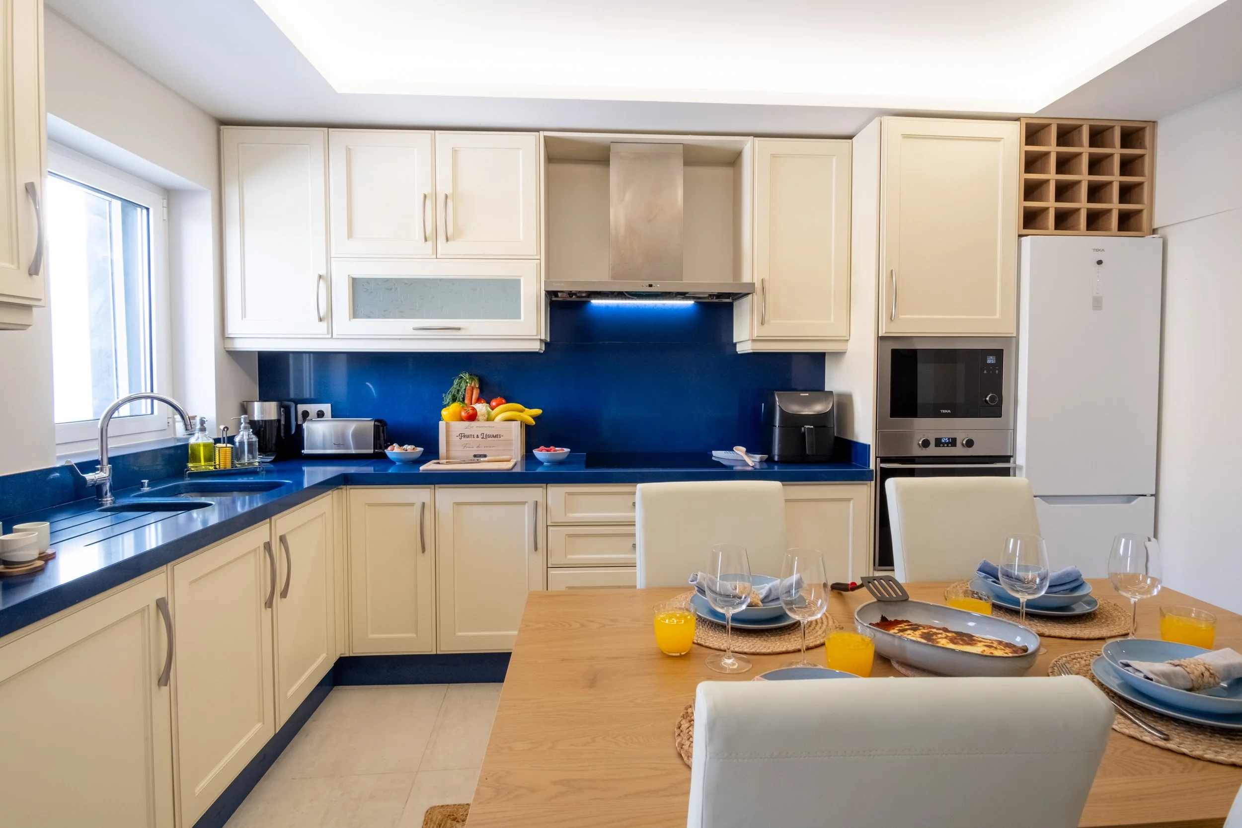 Cozinha moderna com armários brancos, bancada azul, mesa de jantar com pratos, copos de vinho, suco de laranja, cadeiras brancas, e eletrodomésticos de inox, incluindo geladeira, micro-ondas e forno.