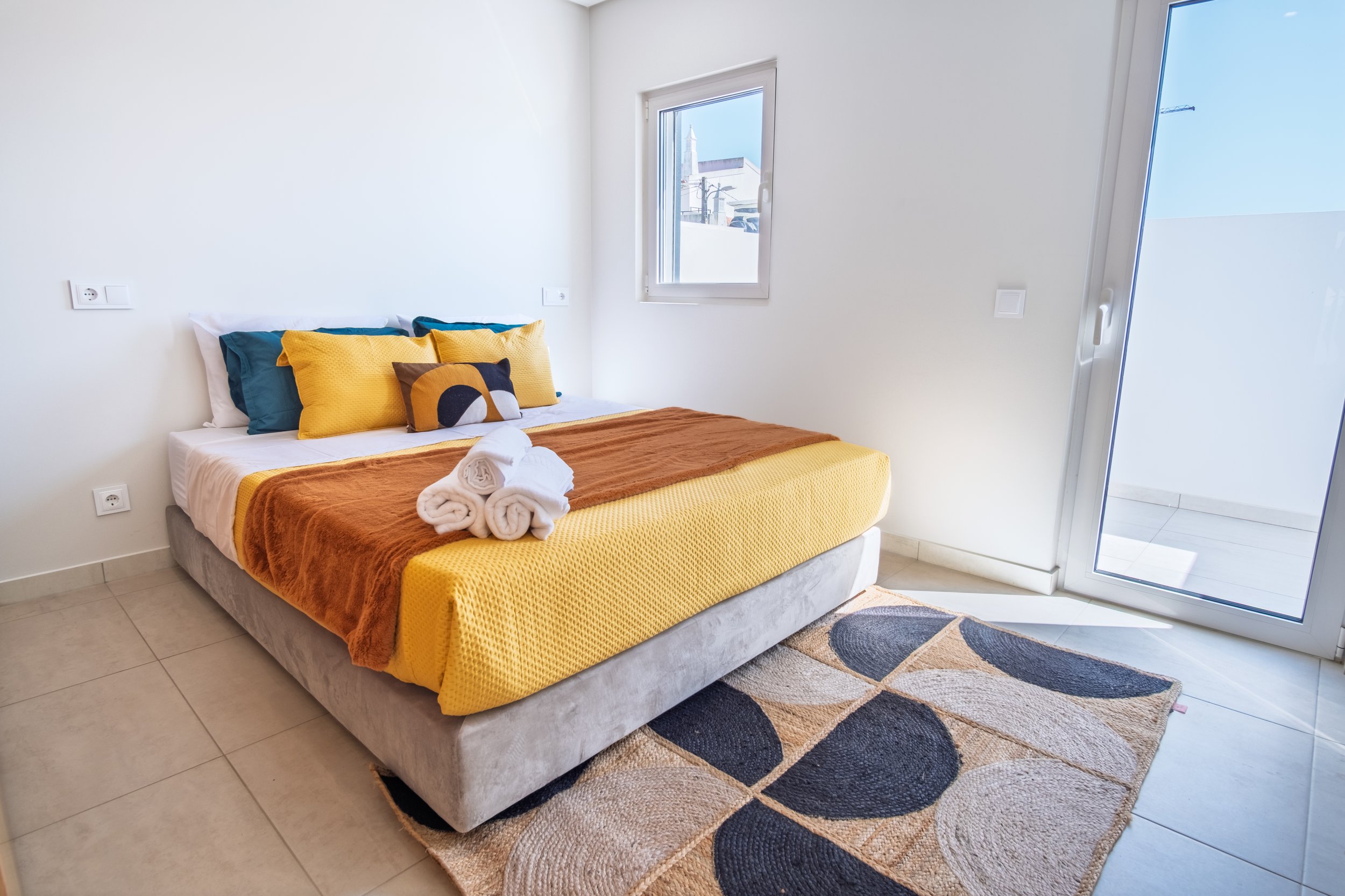 Quarto com cama de casal, roupas de cama coloridas e toalhas dobradas, próximo a uma janela e porta de vidro que leva a uma varanda.