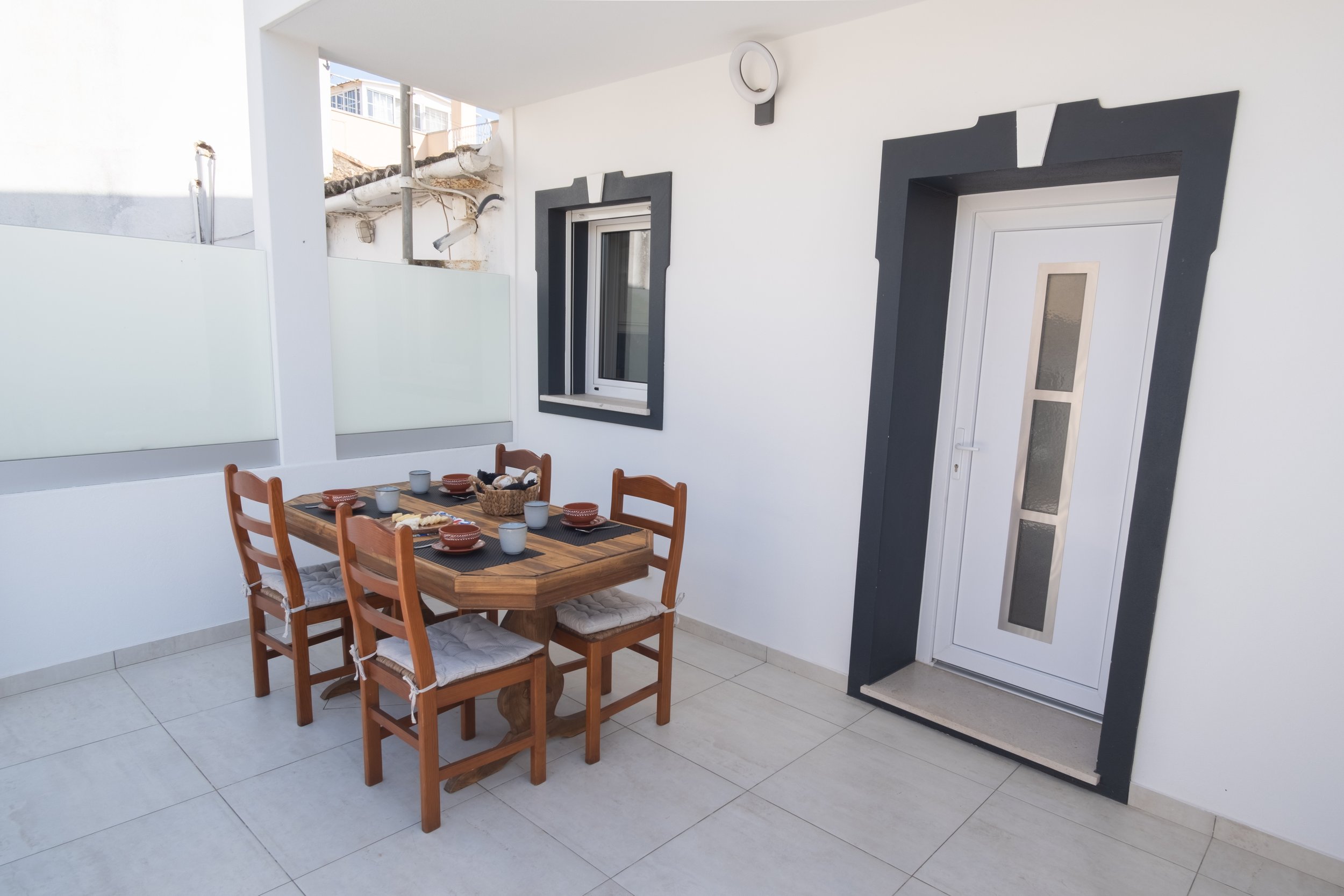 Varanda com mesa de jantar de madeira, quatro cadeiras com assentos de pano, alimentos e utensílios, parede branca com janela e porta branca decorada com moldura escura