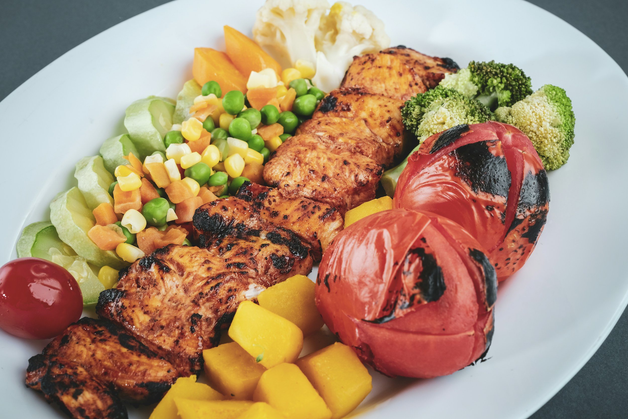 Grilled salmon fillet with charred tomatoes, broccoli, yellow squash, cauliflower, mixed vegetables including corn, peas, and carrots, and a cherry tomato on a white plate.