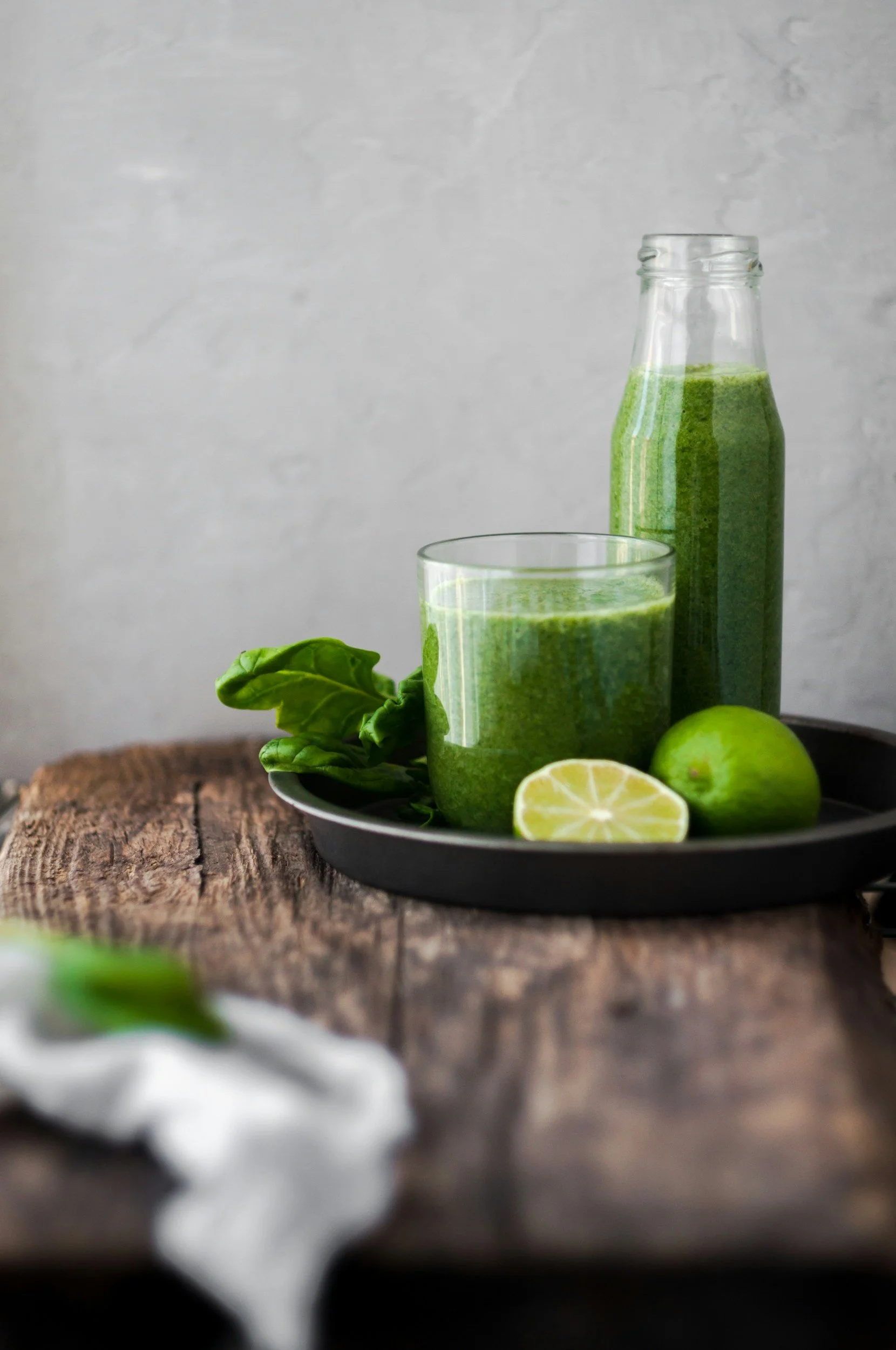A glass of green smoothie, a bottle of green smoothie, a whole lime, a lime half, and fresh basil on a black tray on a rustic wooden surface.