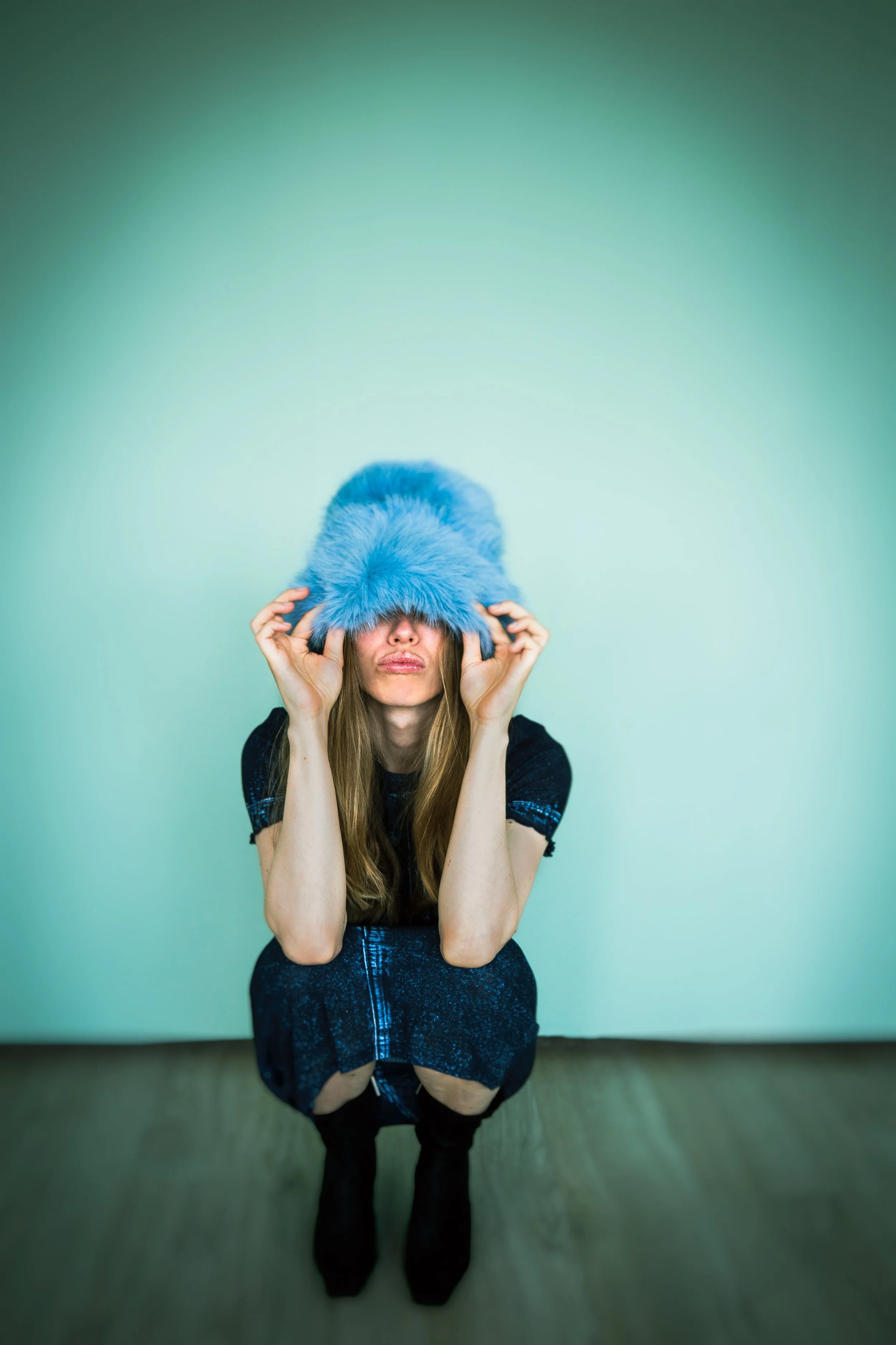 Frau mit langem Haar trägt einen großen, flauschigen blauen Hut, verdeckt ihr Gesicht, sitzt auf dem Boden vor einer hellen Wand.