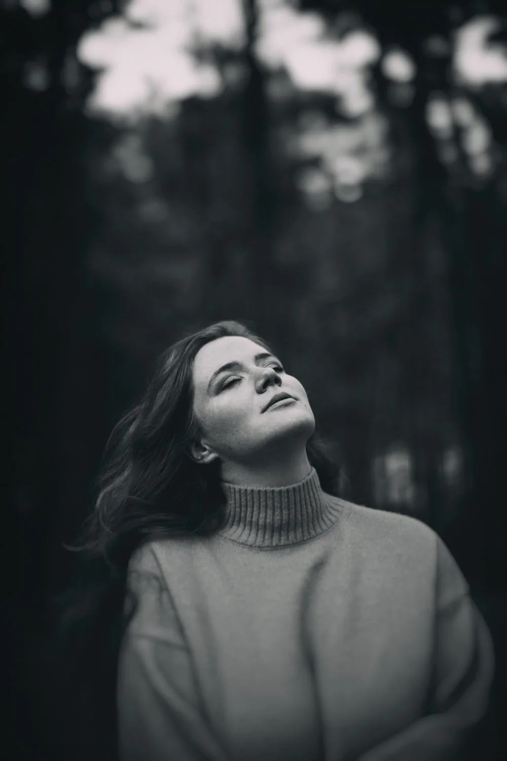Schwarz-weiß-Foto einer Frau mit geschlossenen Augen, die den Kopf leicht nach oben hebt, im Freien vor Bäumen.
