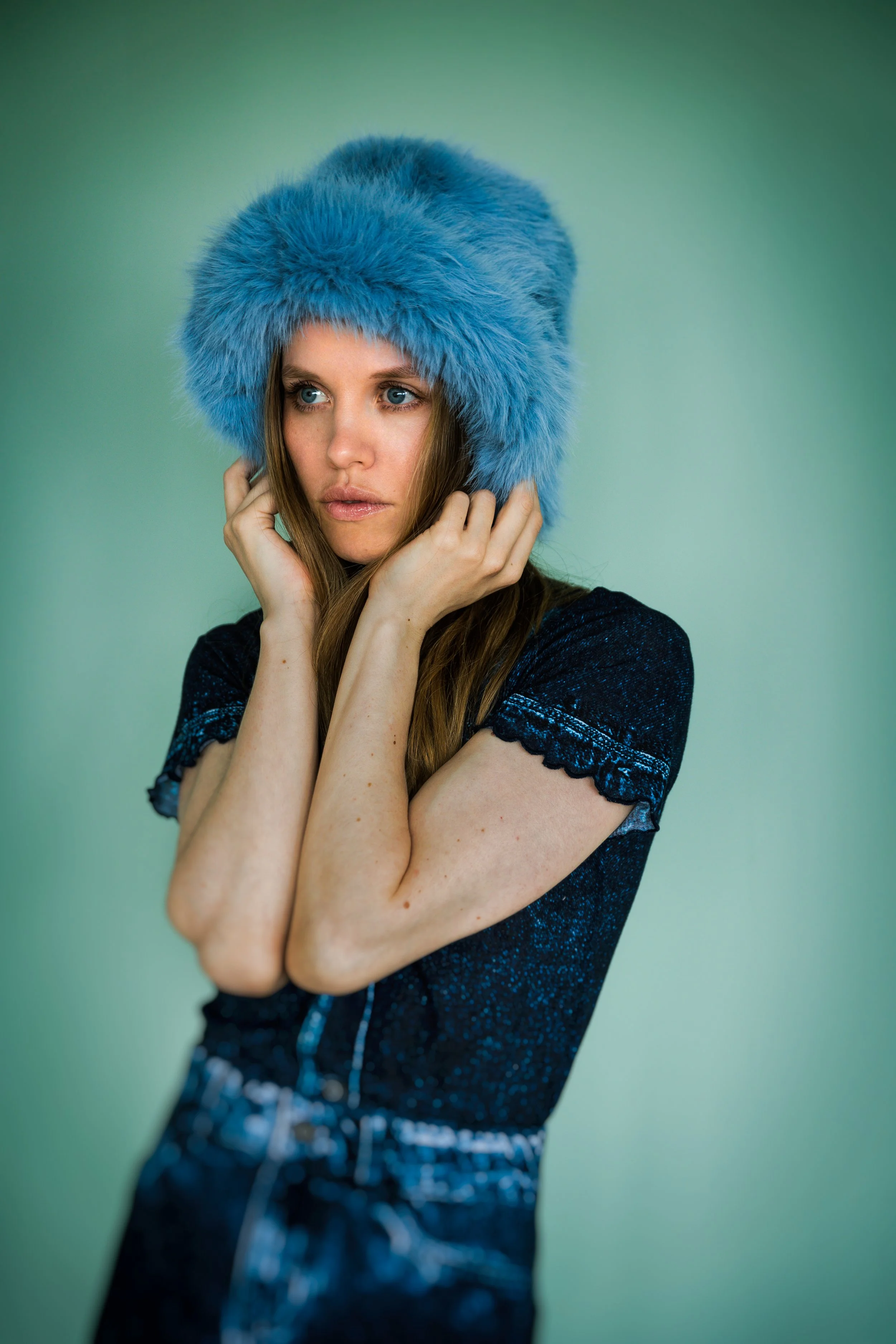 Junge Frau mit langem Haar trägt eine große, blaue, pelzige Mütze und ein schwarzes Oberteil mit Spitzenärmeln vor einem hellgrünen Hintergrund.