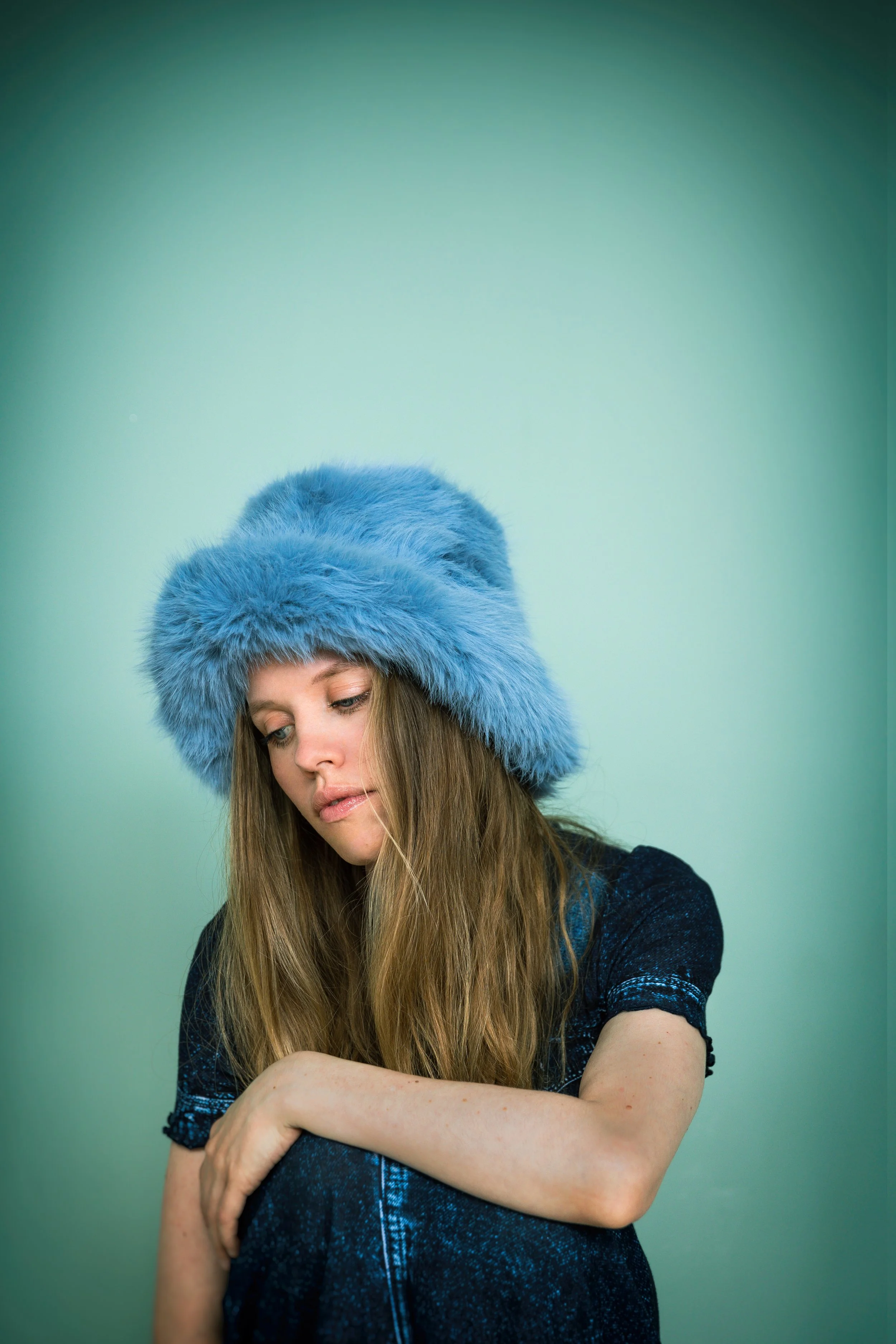 Junge Frau mit langem braunen Haar trägt eine große, flauschige blaue Mütze und hat einen nachdenklichen Ausdruck, sitzt vor einem hellgrünen Hintergrund.