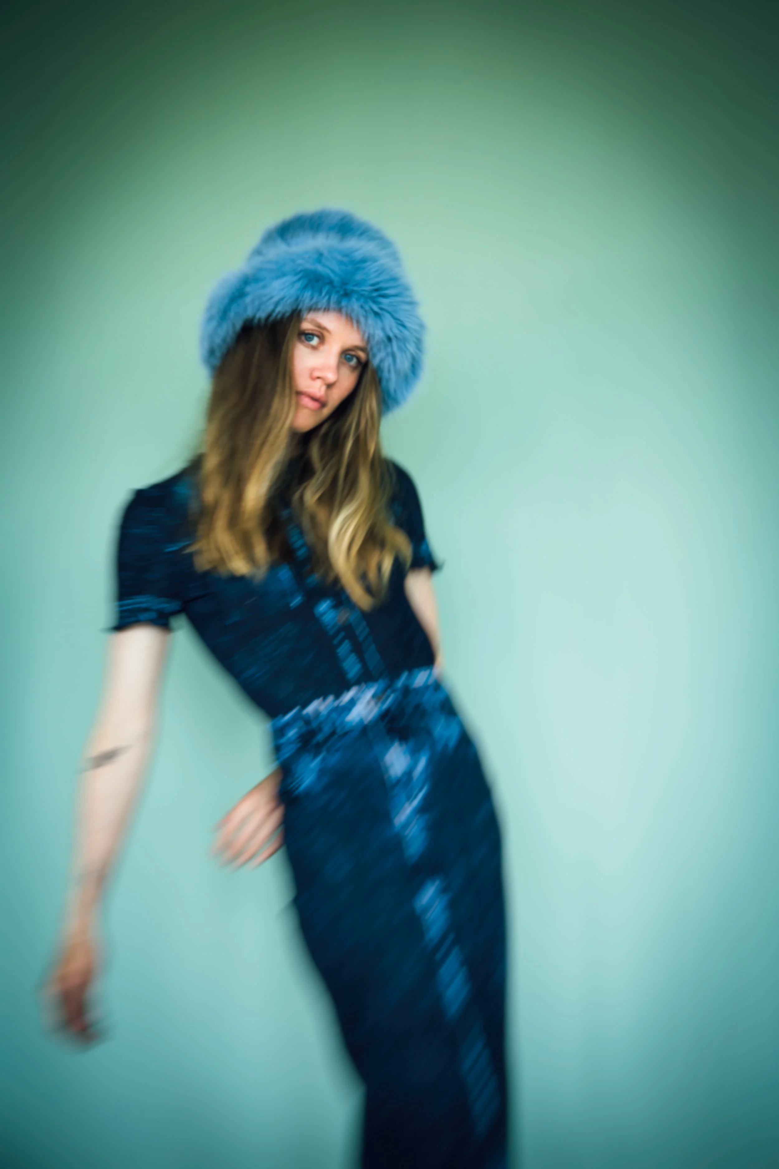 Eine Frau trägt einen blauen, pelzigen Hut und ein blaues Kleid vor einem hellgrünen Hintergrund.