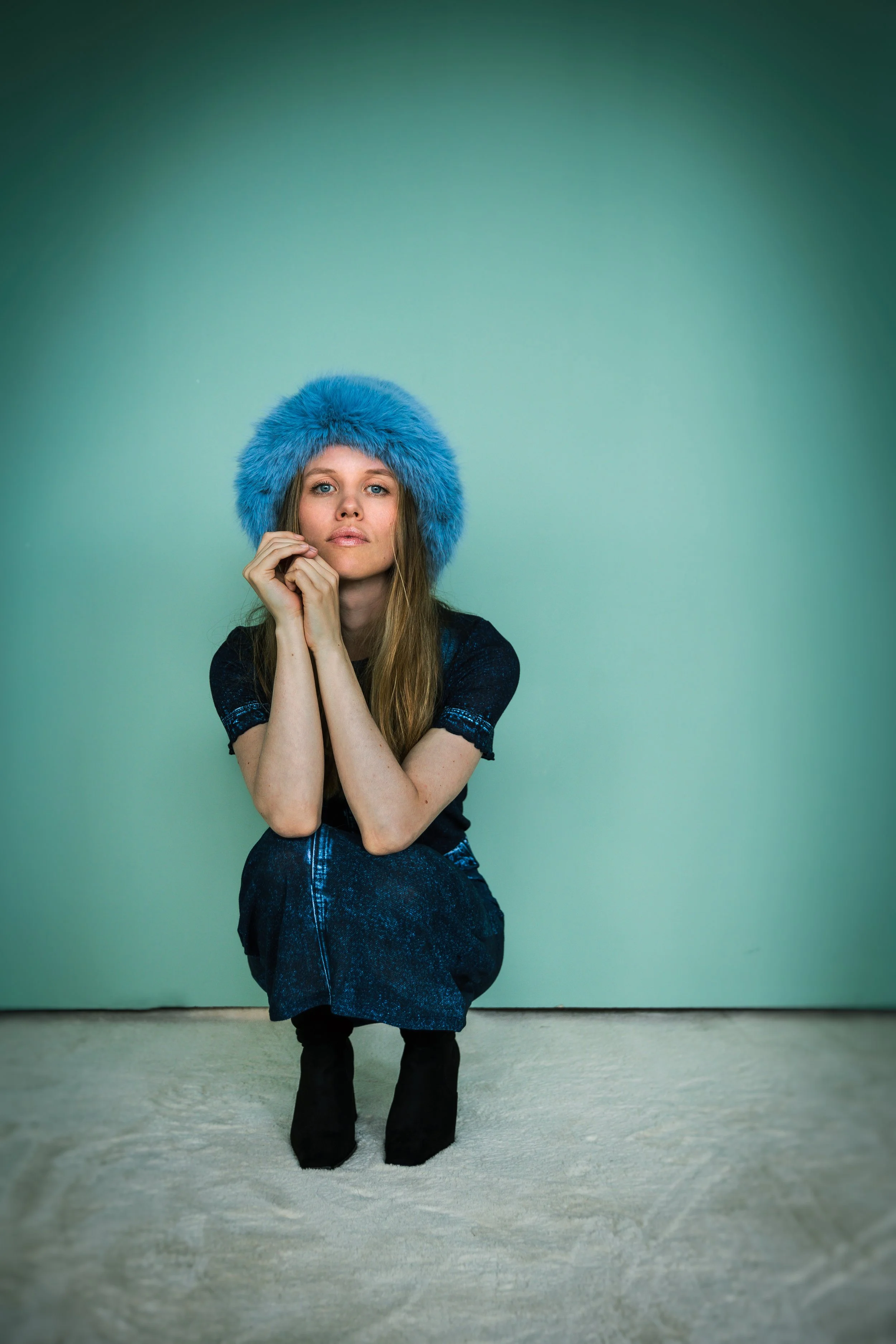 Frau mit blauer Modef zu Hüte, sitzend im Schneidersitz auf grauem Boden vor grünlicher Wand.