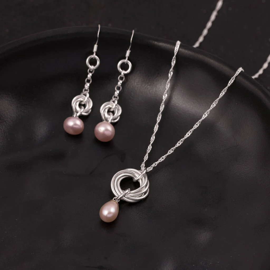 Silver jewellery set including a necklace with a pink pearl pendant and matching earrings with pink pearl accents, displayed on a black surface.