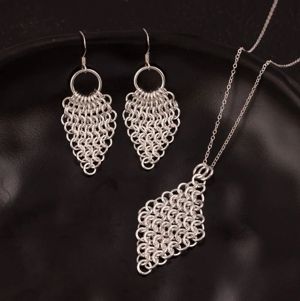 A set of silver jewellery, including a pair of chainmaille earrings and a matching chainmaille necklace, displayed on a black surface.