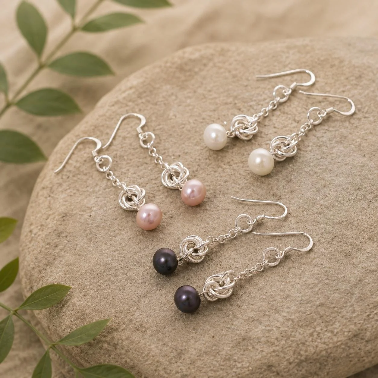 Set of three silver chain earrings with pearl and bead pendants on a beige stone, surrounded by green leaves.