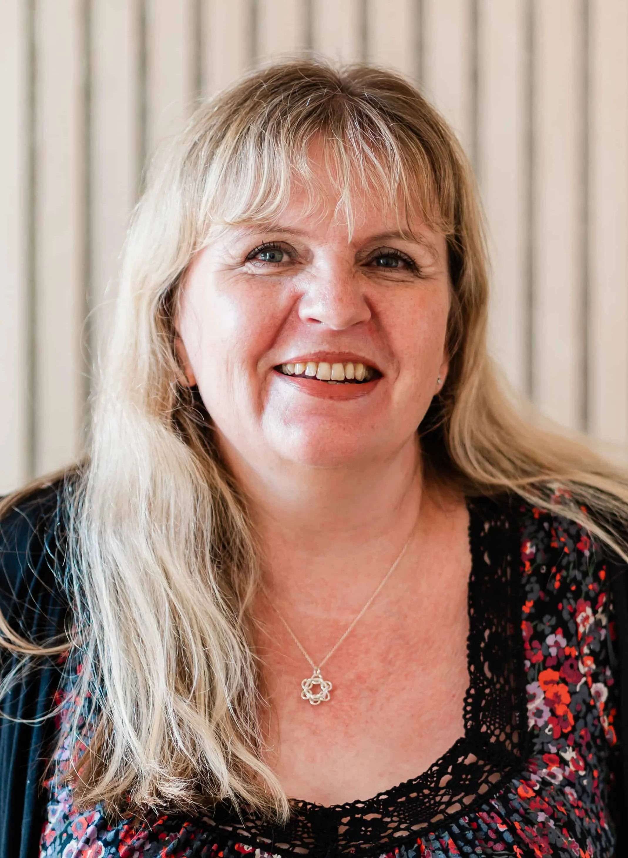 Close-up of a smiling middle-aged woman with long blonde hair, wearing a black dress with red and purple floral pattern, a silver necklace, and a silver flower-shaped pendant.