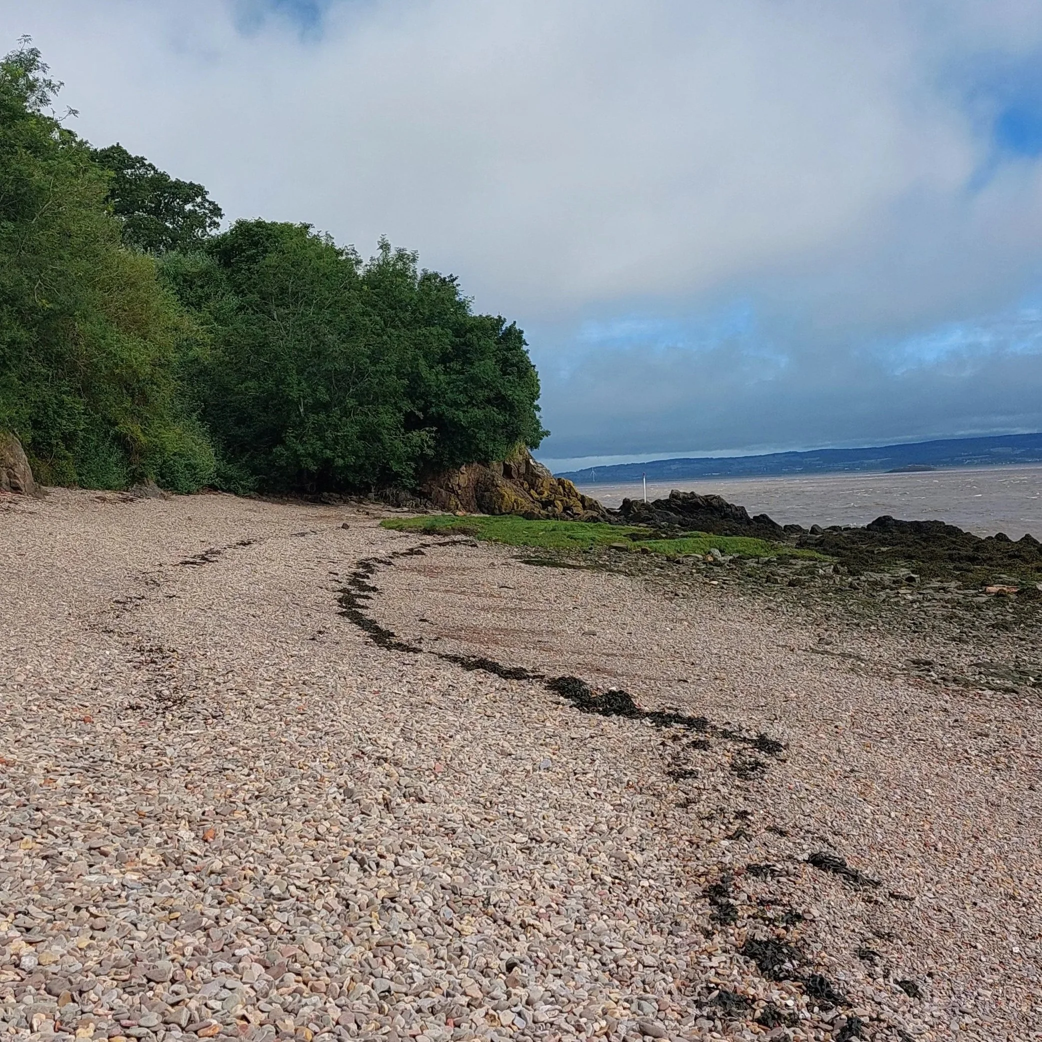 A rocky beach with pebbles and stones leading to water, backed by lush green trees on a hillside and a cloudy sky above with some blue patches.