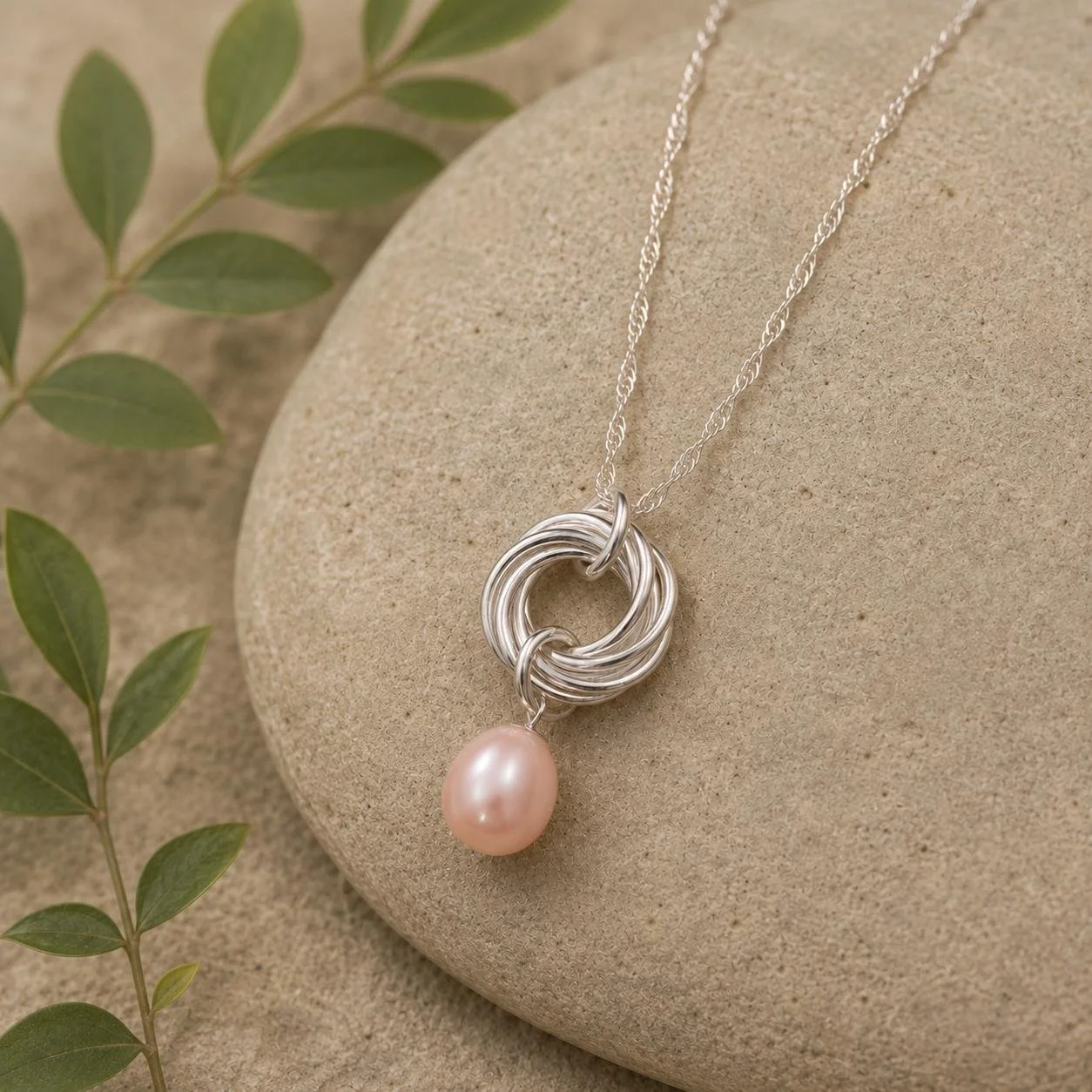 Silver necklace with a knot pendant and pink pearl, displayed on a sandy surface with green leaves nearby.