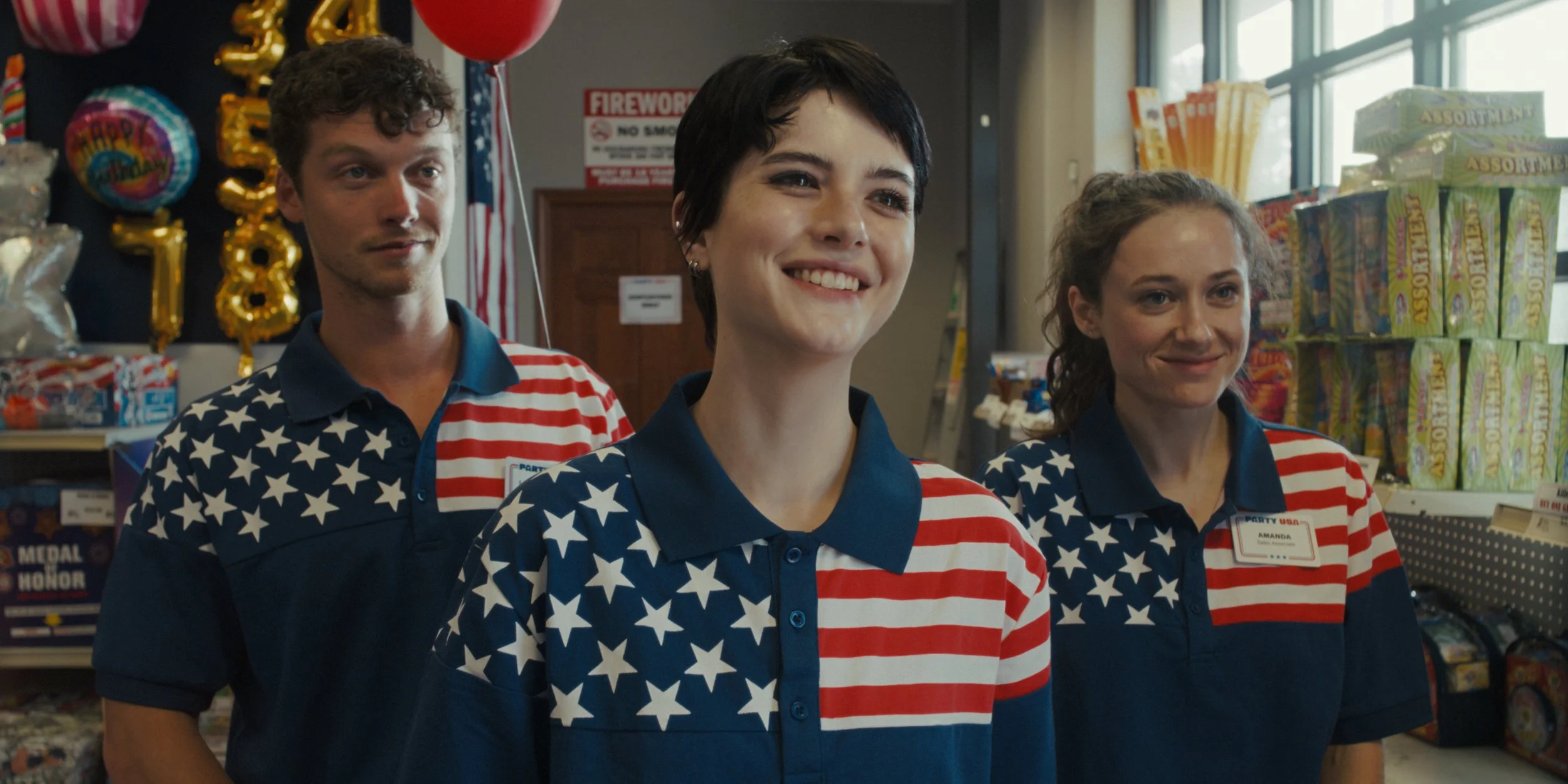 Three store employees with American flag-themed uniforms, standing inside a store decorated for a celebration with balloons and banners.