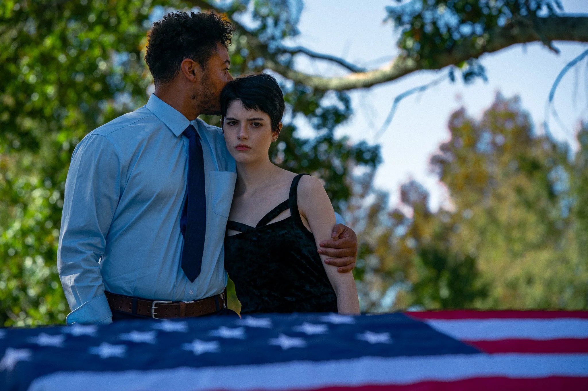 A man with curly hair and a woman with short black hair standing outdoors, with an American flag draped in front of them. The man is wearing a light blue shirt and a dark tie, embracing the woman who has a somber expression and is dressed in a black 