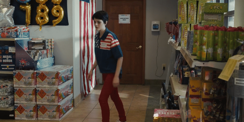 Ainsley Seiger in a patriotic sweater and red pants walking inside a store decorated with gold balloons forming the number 189, with shelves stocked with fireworks and other items.