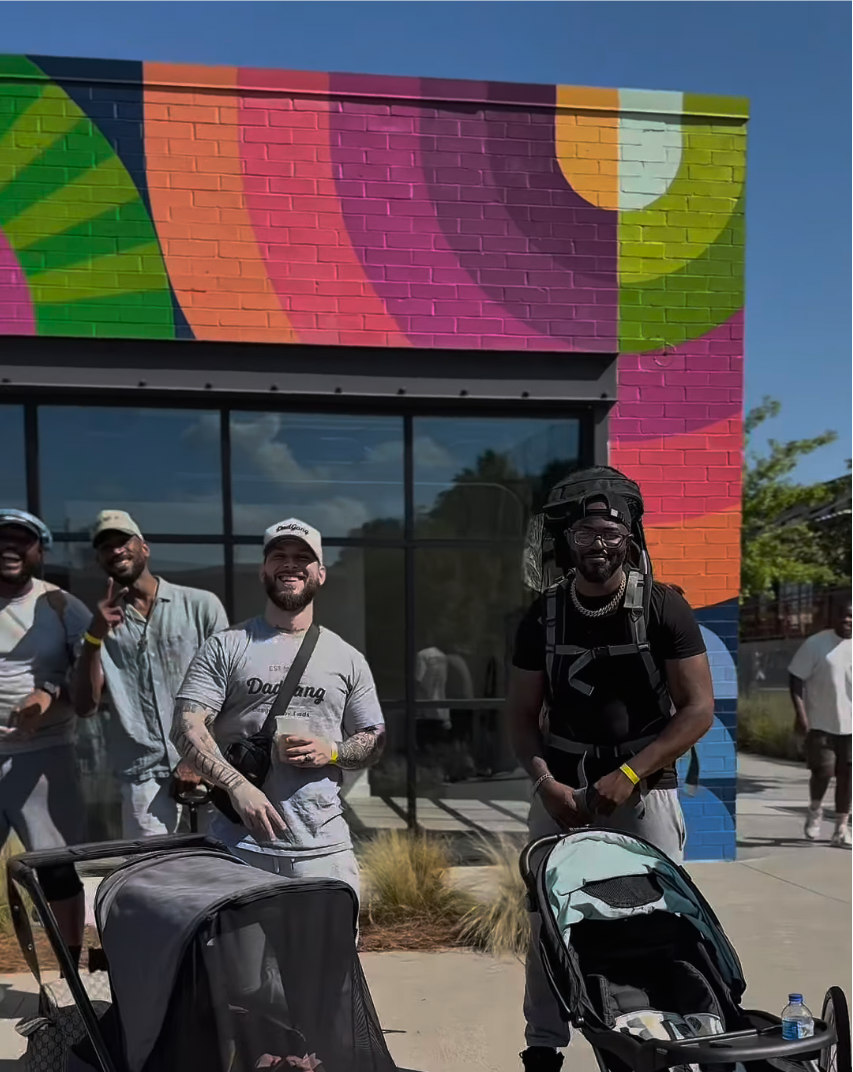 A group of men standing in front of a colorful, mural-painted brick building with large windows. Two men are pushing strollers, one with drinks, and others are smiling and posing for the photo.
