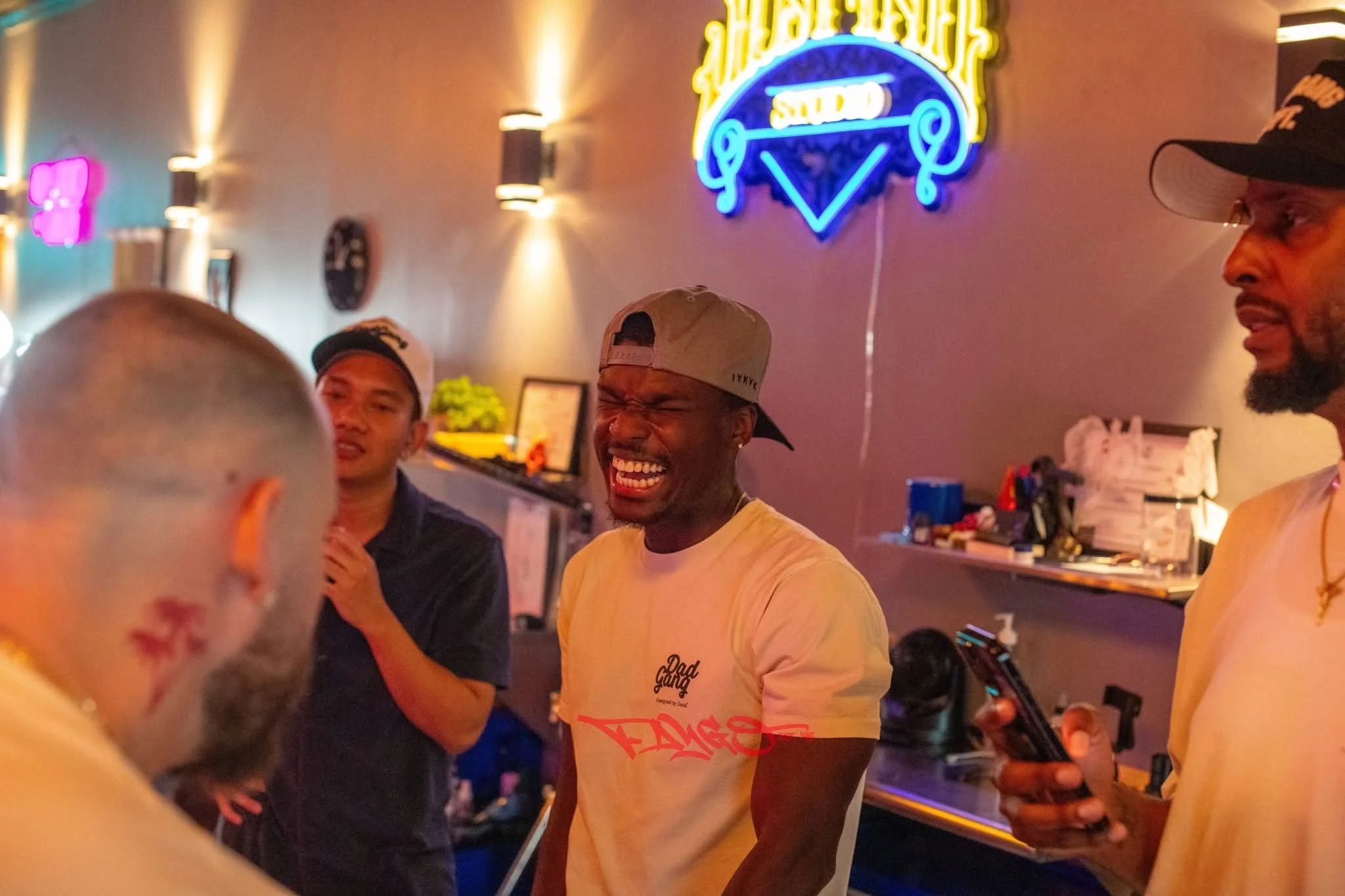 Group of four men at a bar or restaurant, with neon signs on the wall behind them, one of the men is laughing with eyes closed.
