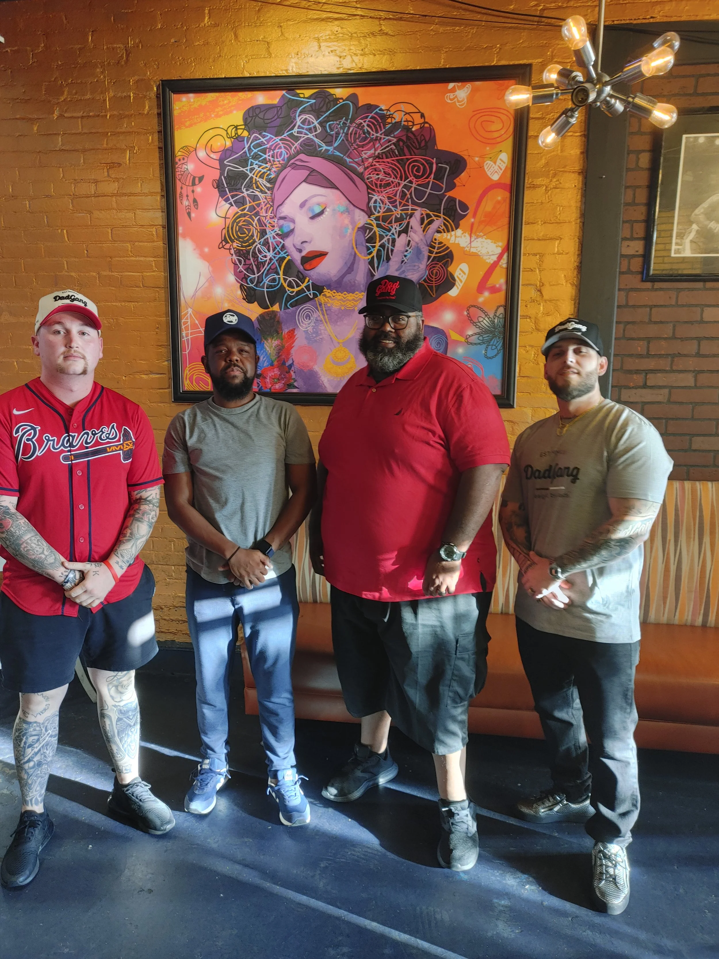 Four men standing in front of an orange brick wall with a colorful framed artwork of a woman with curly hair and vibrant makeup. One man wears a red Atlanta Braves jersey, another a gray t-shirt, the third a red polo shirt, and the last a gray t-shir