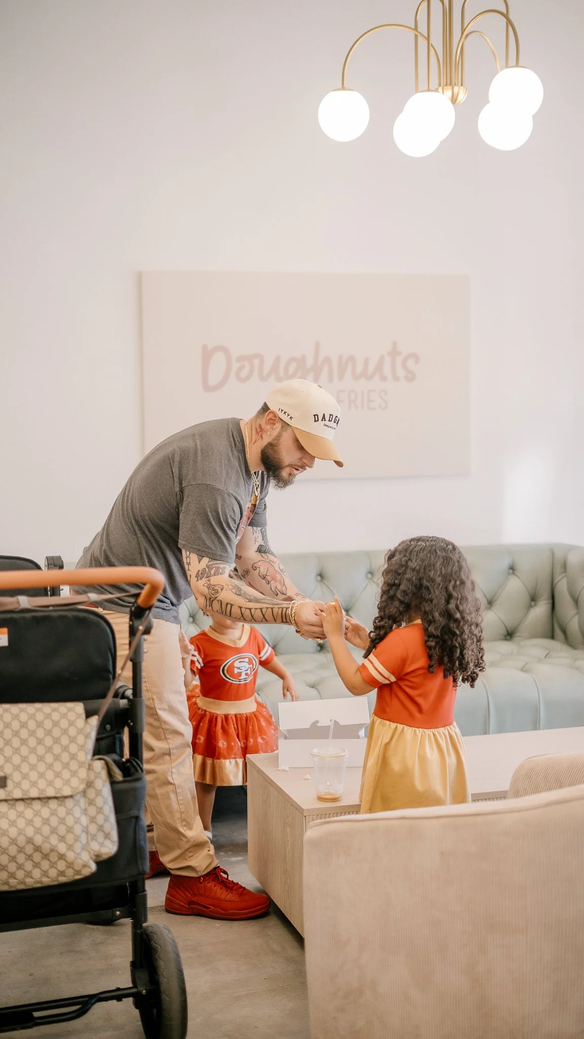 A man with tattoos, wearing a gray T-shirt, a white and tan cap, red sneakers, and beige pants, is handing a piece of food to a young girl with curly hair, wearing an orange and yellow dress, in a casual café or bakery setting. There is a baby girl i