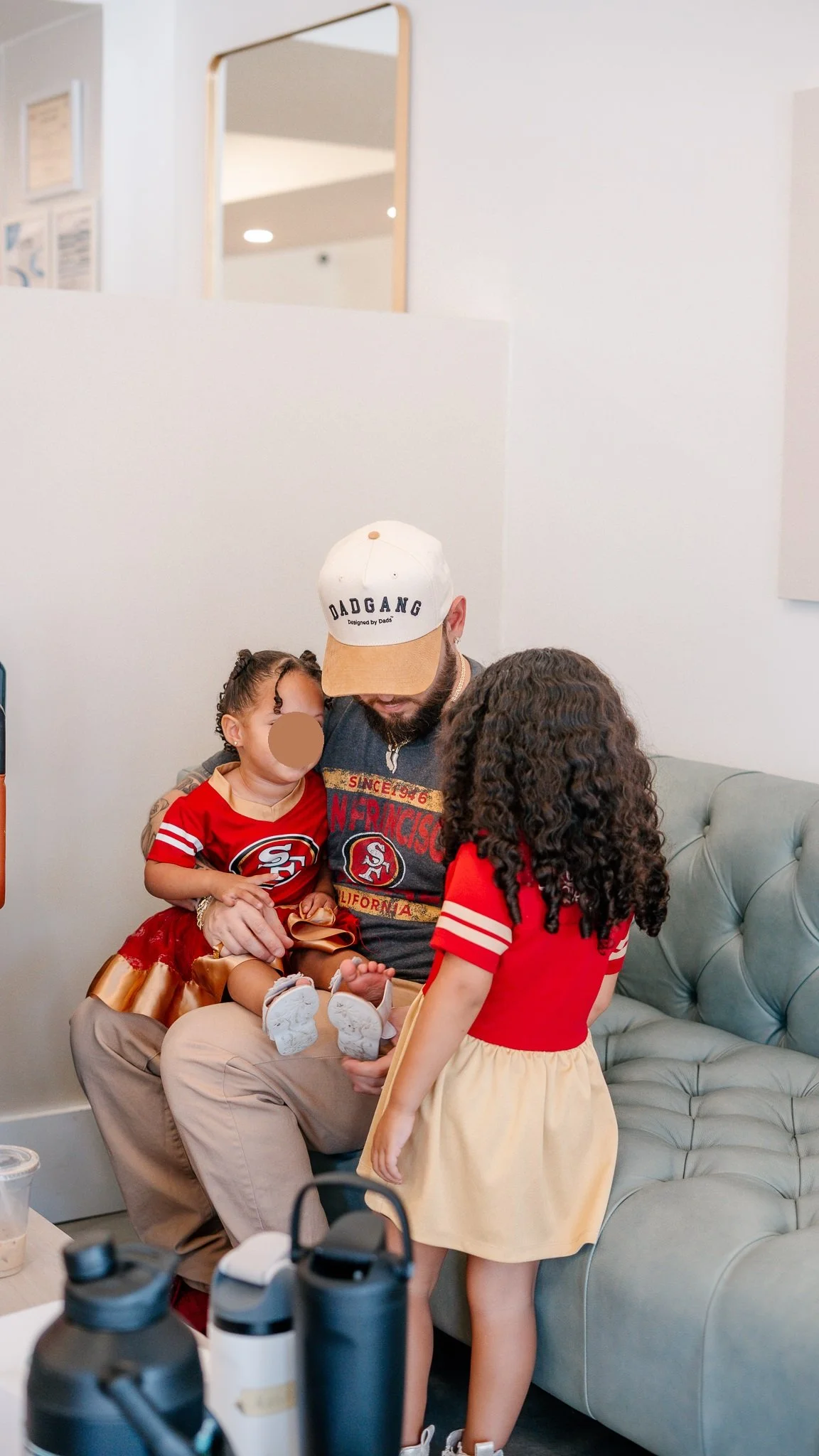 A man wearing a San Francisco 49ers shirt, a white and tan cap, and beige pants sits on a tan tufted couch holding a young girl dressed in a red and gold 49ers outfit. A girl with curly hair in a red top and beige skirt stands nearby, facing the man 