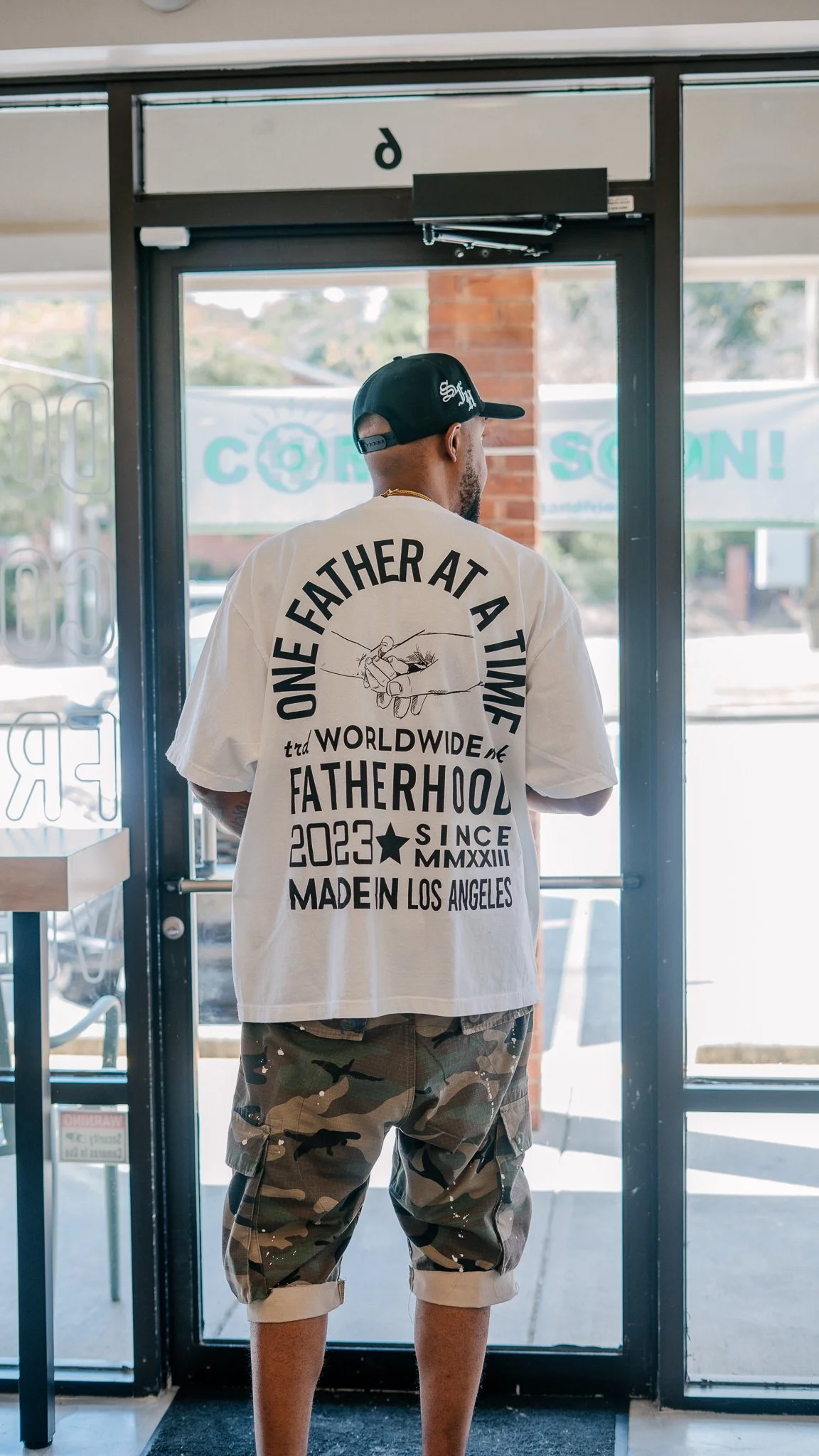 A man stands with his back to the camera at a doorway, wearing a white graphic T-shirt and camouflage shorts, with a black cap. The T-shirt has text and illustrations referencing fatherhood.