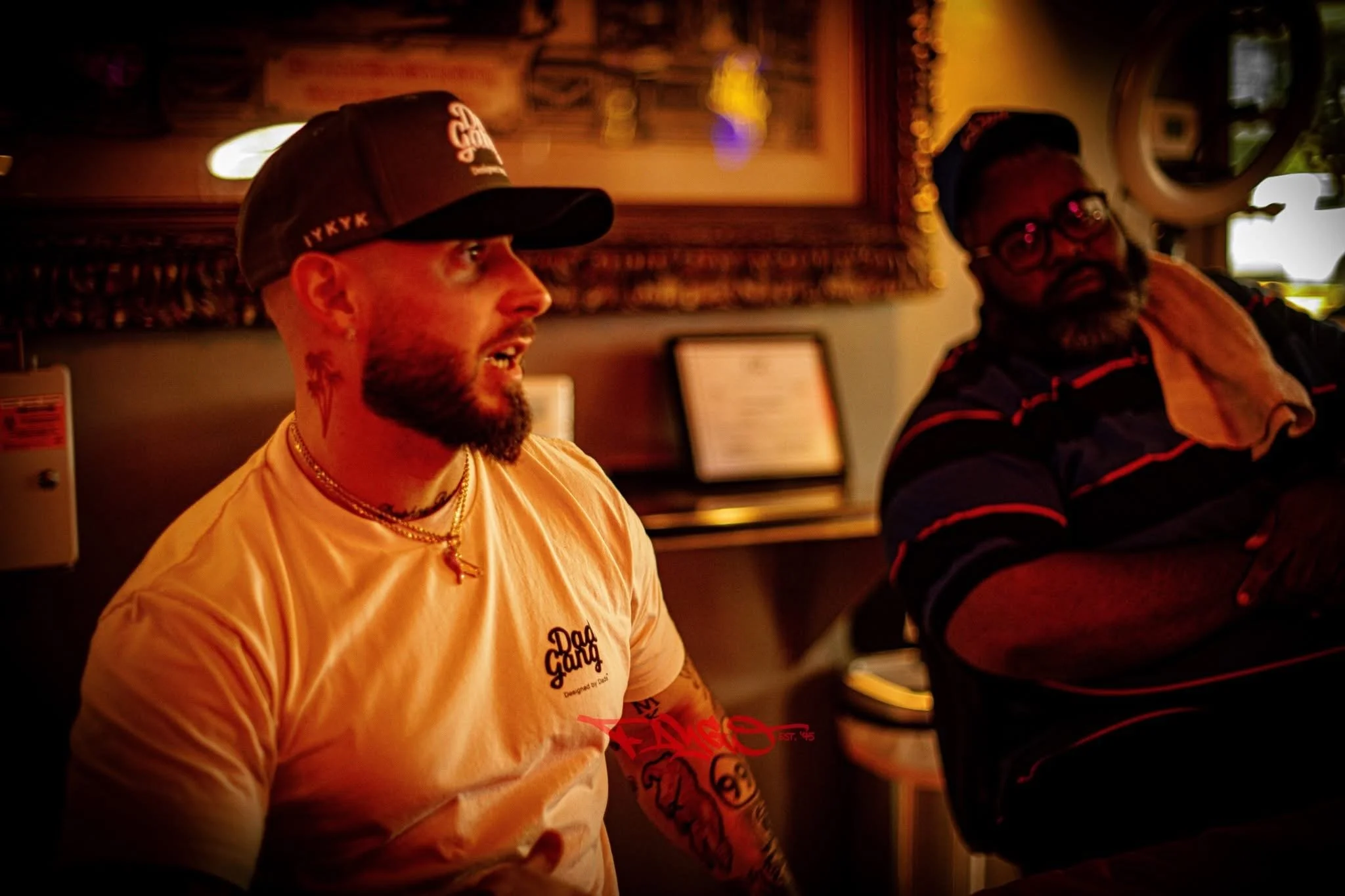 Two men sitting indoors in a dimly lit environment, engaged in conversation. The man on the left is wearing a cap, a light-colored t-shirt, and gold jewelry, and has a tattoo on his neck. The man on the right is wearing glasses, a striped shirt, and 