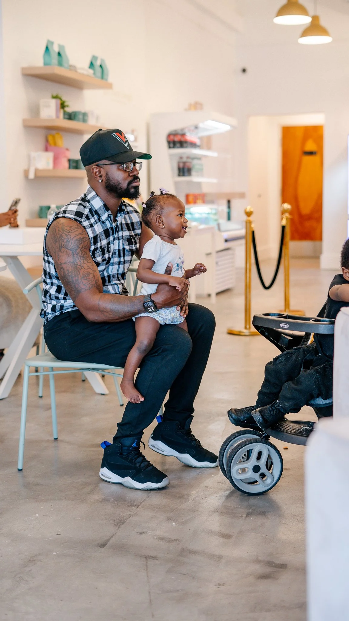 A man with tattoos, wearing glasses, a black cap, a sleeveless checkered shirt, black pants, and black sneakers, is sitting on a chair holding a young girl on his lap. The girl is smiling, wearing a white shirt and shorts. They are inside a brightly 