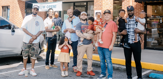Group of adults and children standing outside a storefront, smiling for the photo. Some children are being held by adults, and there are parked cars nearby.