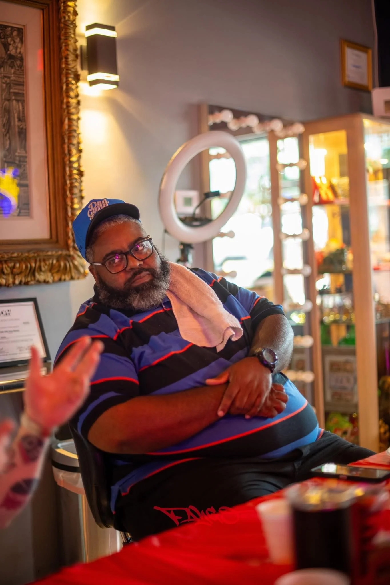 A man with glasses and a beard wearing a blue cap, striped polo shirt, and a towel around his neck sitting at a table in a colorful indoor setting, possibly a restaurant or cafe, with decorative lighting and shelves in the background.