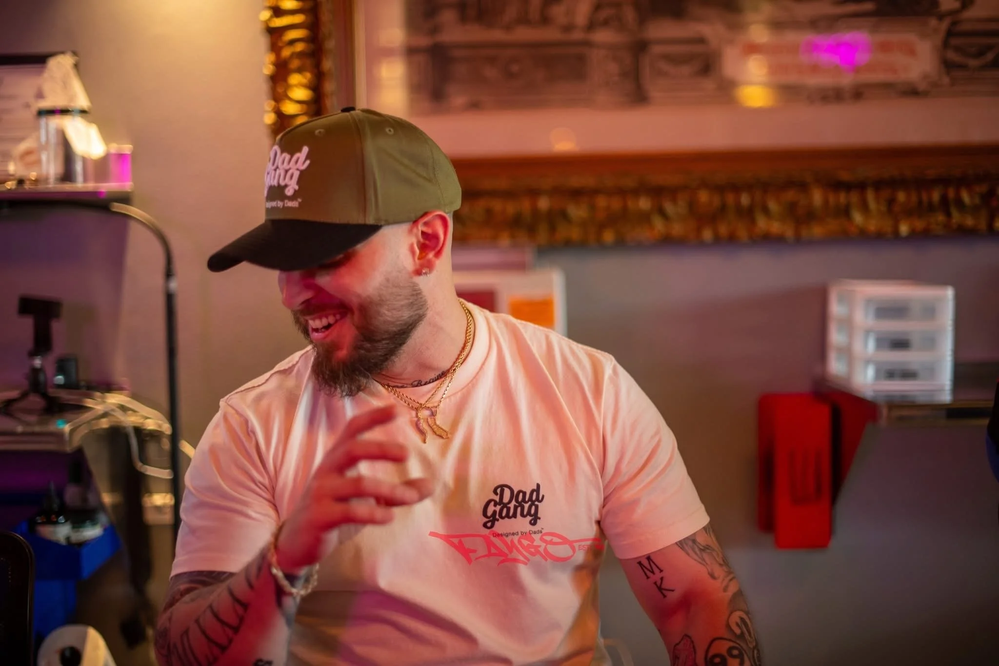 A smiling man with tattoos on his arms, wearing a beige t-shirt and a baseball cap, appears to be enjoying himself in a dimly lit indoor setting.