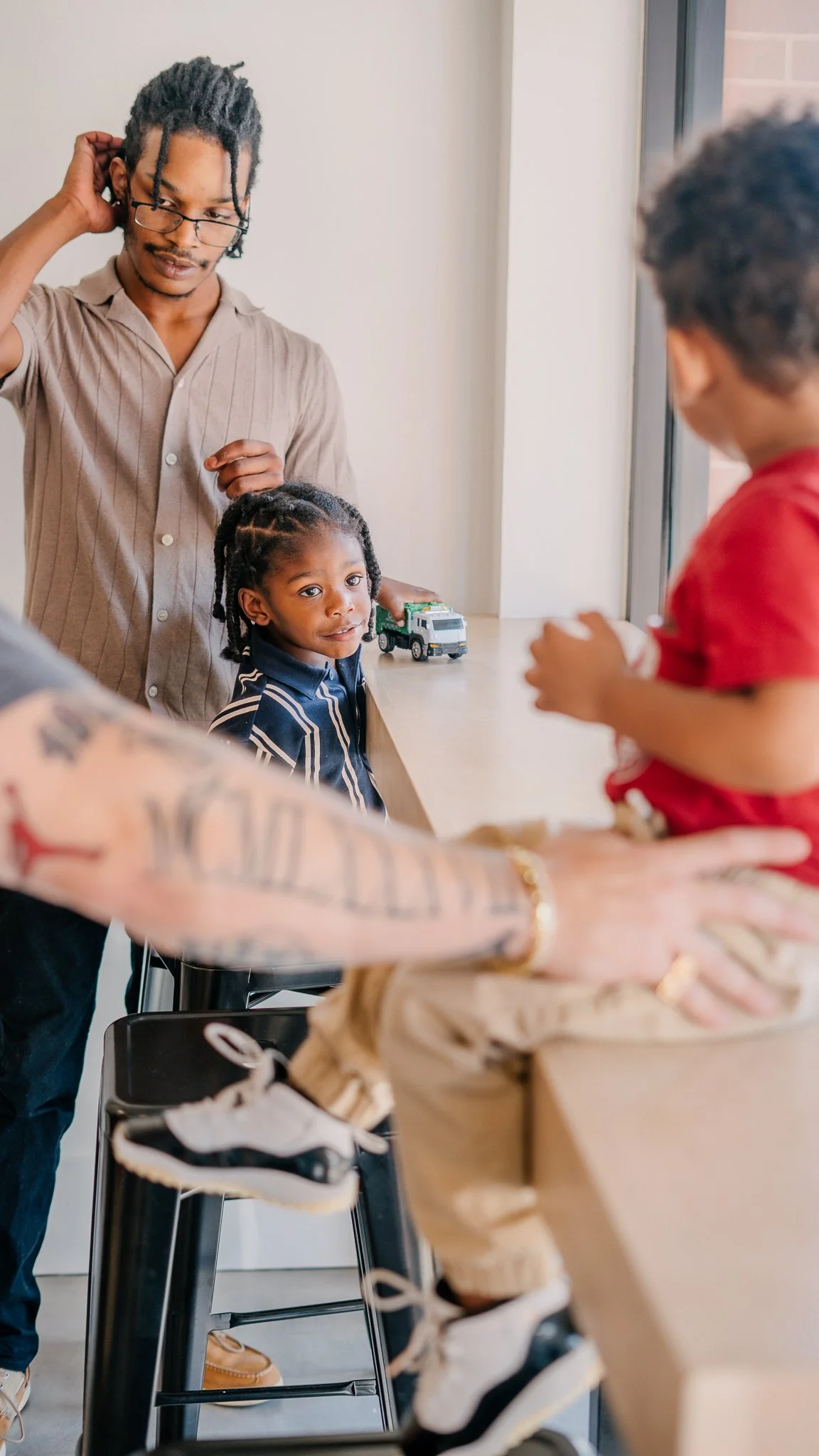 A group of people at a table near a window, including a young girl with braided hair and a person with tattoos on their arm, interacting with a boy in a red shirt. Toys are on the table and a window is in the background.