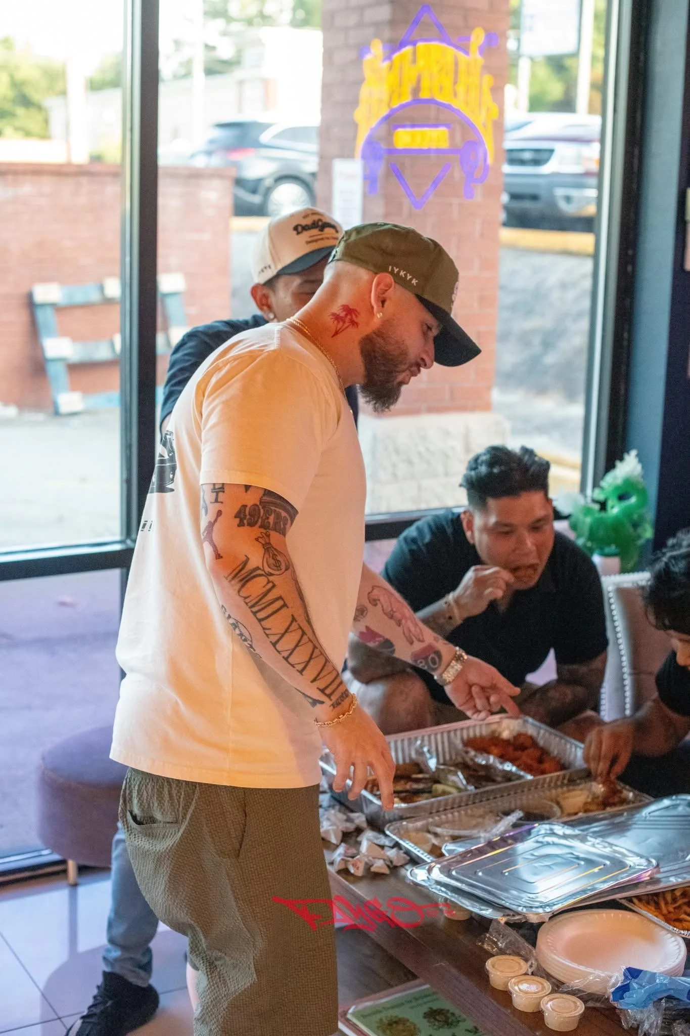 A group of people gathered around a table with food, inside a restaurant with large windows and a brick wall outside.