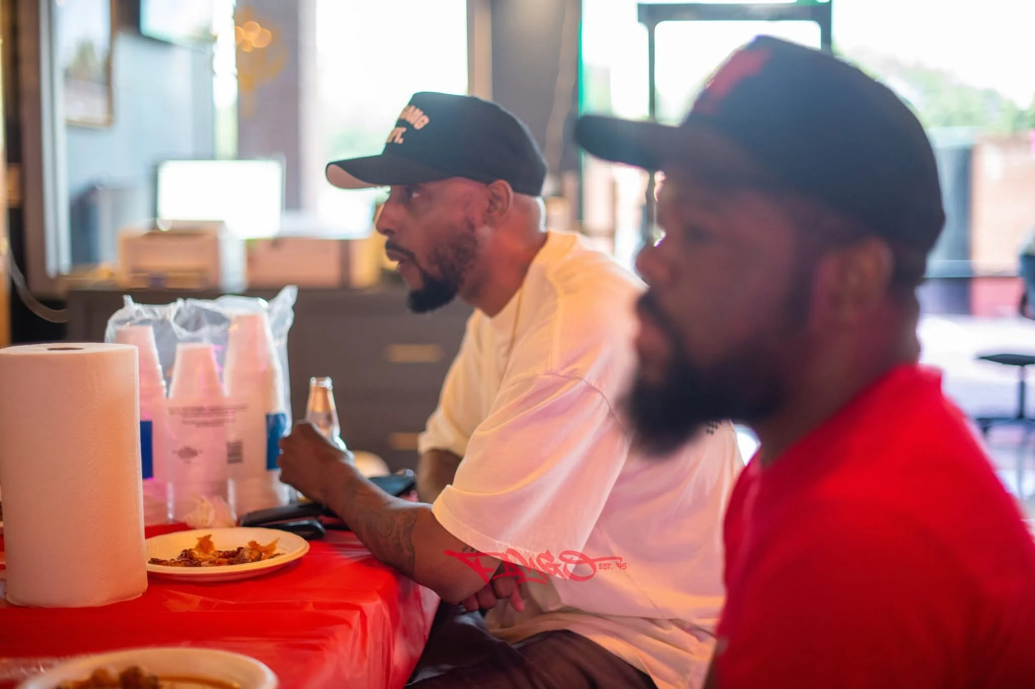 Two men sitting at a table in a restaurant or cafe, wearing caps; one in a white shirt, the other in red, with food, paper towels, and bottles on the table.