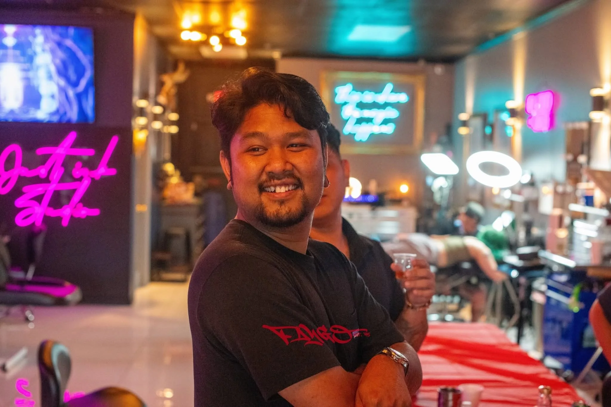 A smiling man with dark hair, beard, and earrings, standing in a bar with neon signs and colorful lighting.