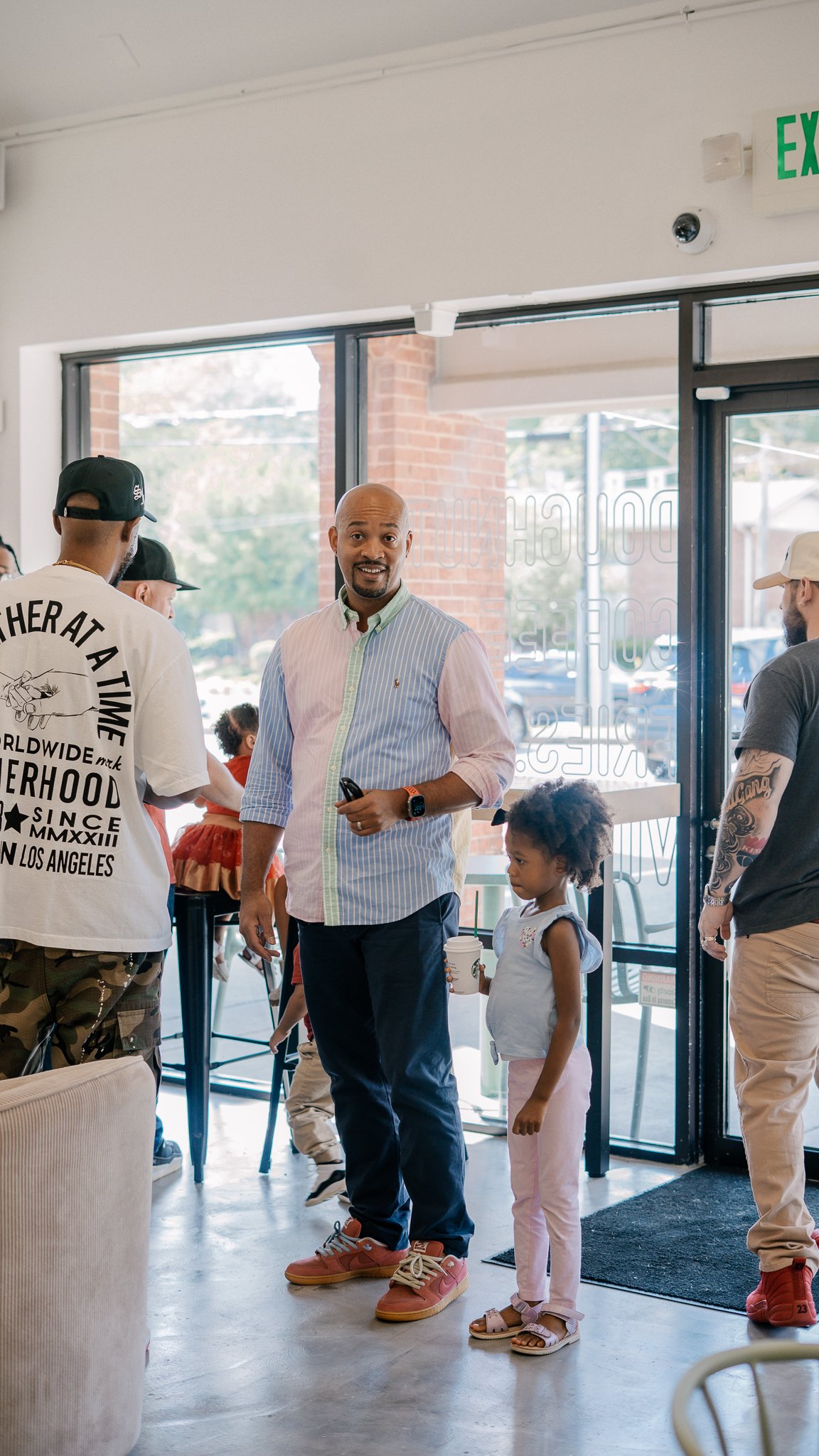 People inside a coffee shop near the entrance, with one man holding a phone and a young girl holding a coffee cup, all engaging in conversation.