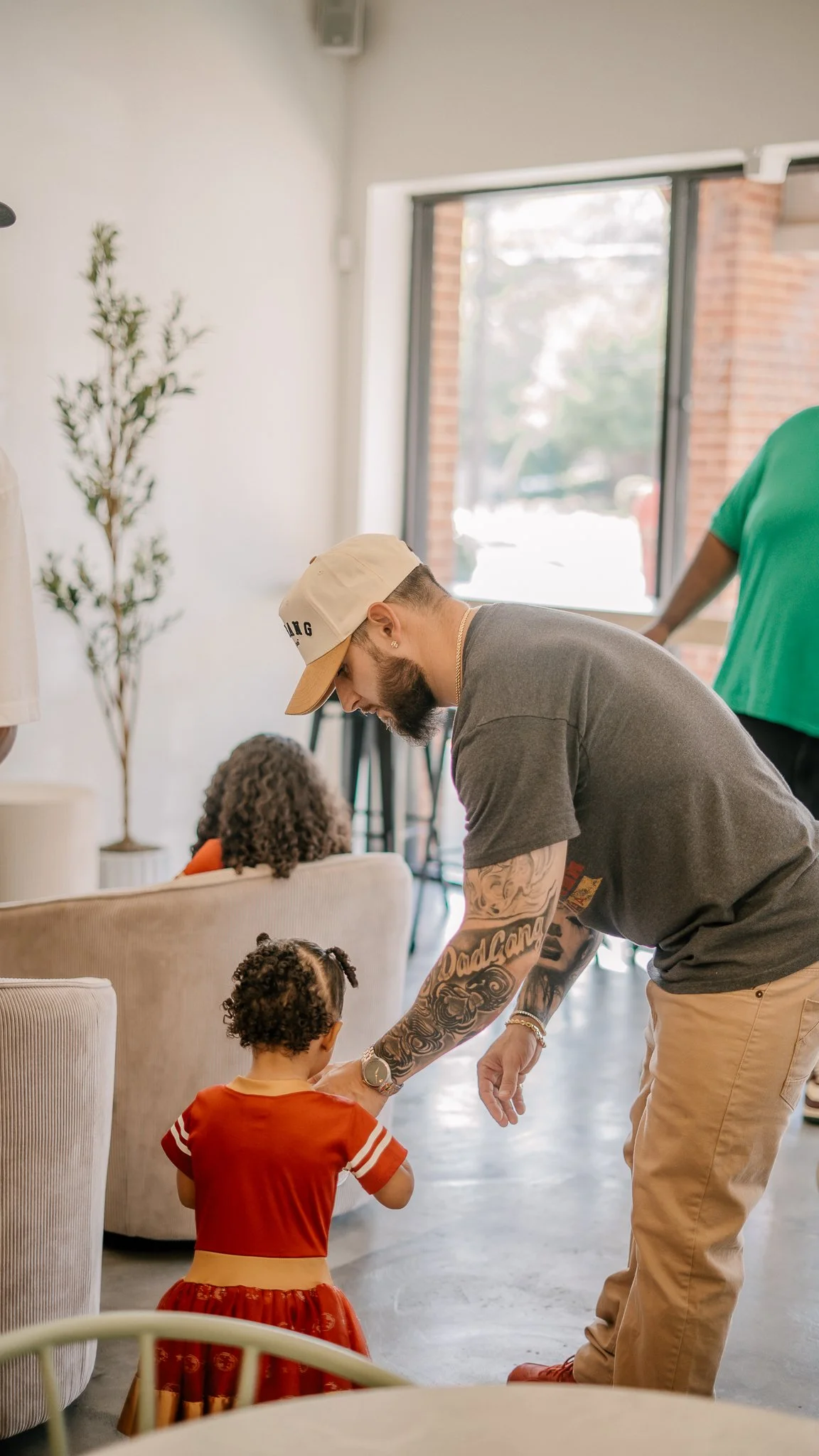 A man with tattoos, wearing a beige baseball cap and gray T-shirt, is bending down to interact with a young girl in an orange dress in a bright, modern room with large windows and minimal decor.