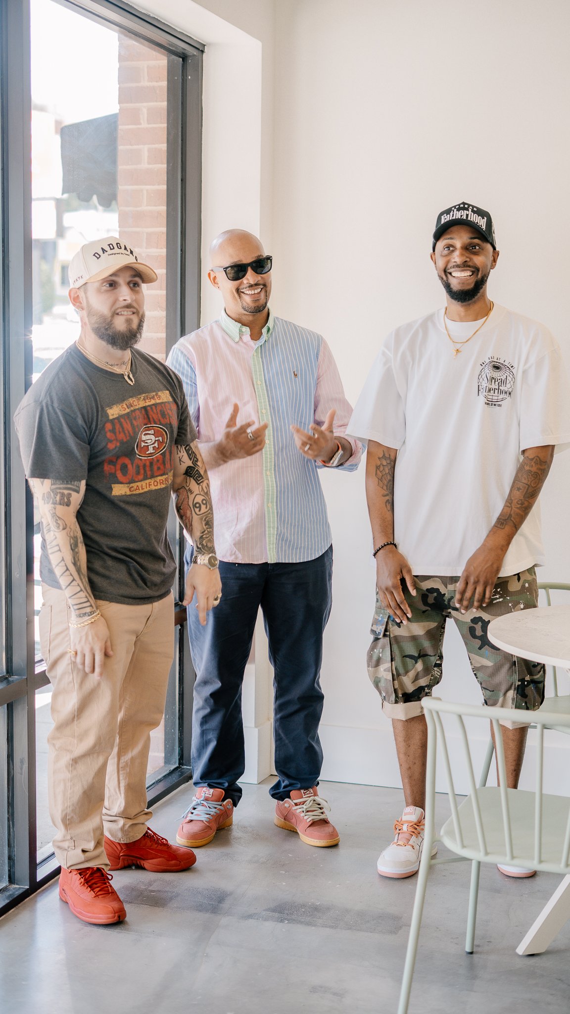 Three men standing near a window inside a cafe, smiling and engaging in conversation.