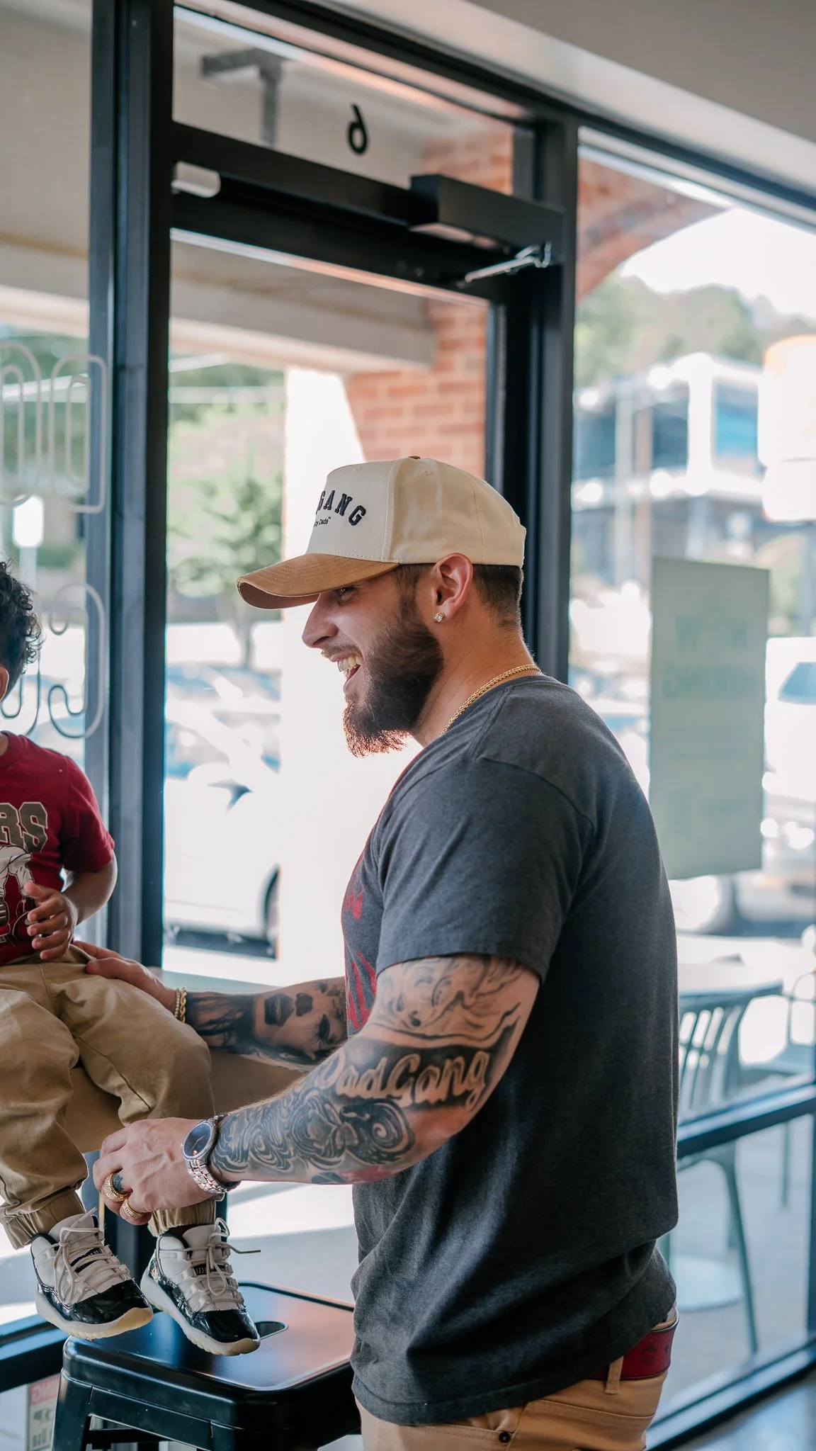 A man with tattoos on his arms, wearing a beige cap, a dark t-shirt, and tan pants, is smiling and holding a young boy sitting on a counter inside a store or cafe.
