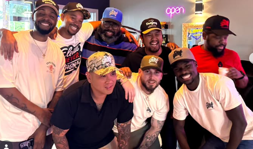 Group of seven smiling men, some wearing baseball caps and t-shirts, posing together in an indoor space with neon lights that spell 'open' in the background.