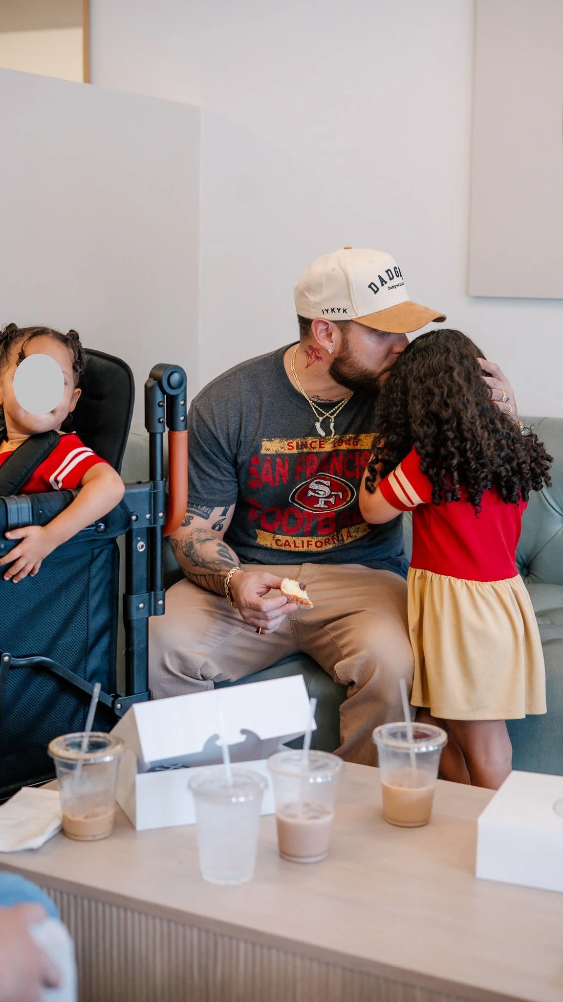 A man with tattoos and a child kissing in a crowded room, with another child in a stroller and drinks on a table.