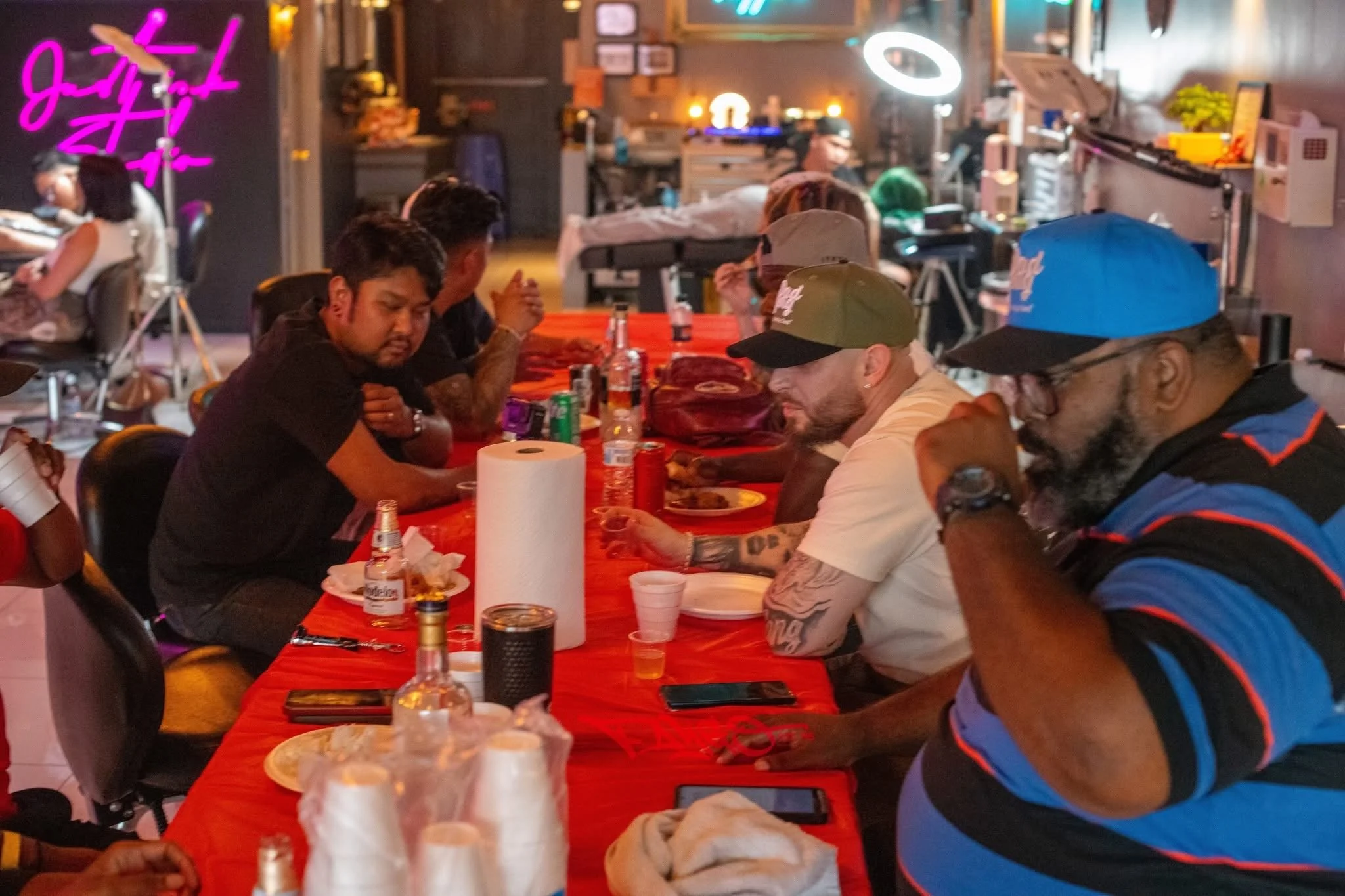 People sitting at a long table in a dimly lit restaurant or bar, some with food and drinks, engaging in conversation. Neon signs and various decorations are visible in the background.