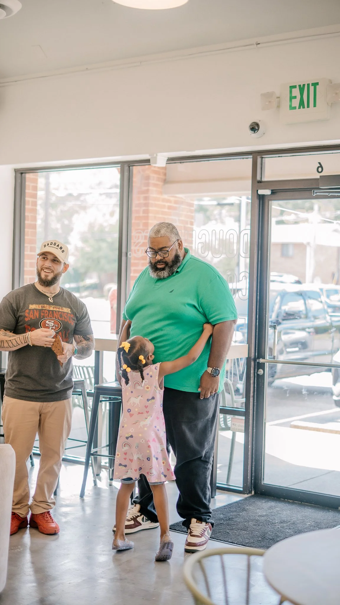 A girl hugging a man at the entrance of a cafe while another man stands nearby with a slice of pizza. The girl has braids with yellow hair ties and is wearing a pink dress with rainbow and unicorn patterns. The man being hugged is wearing a green pol