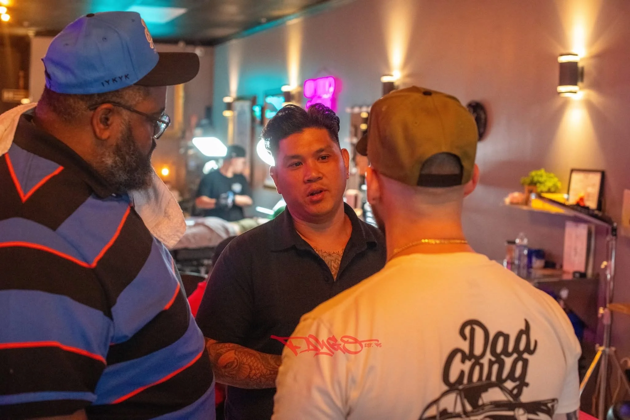 Three men engaged in conversation inside a dimly lit room with colorful lights and neon signs on the wall.